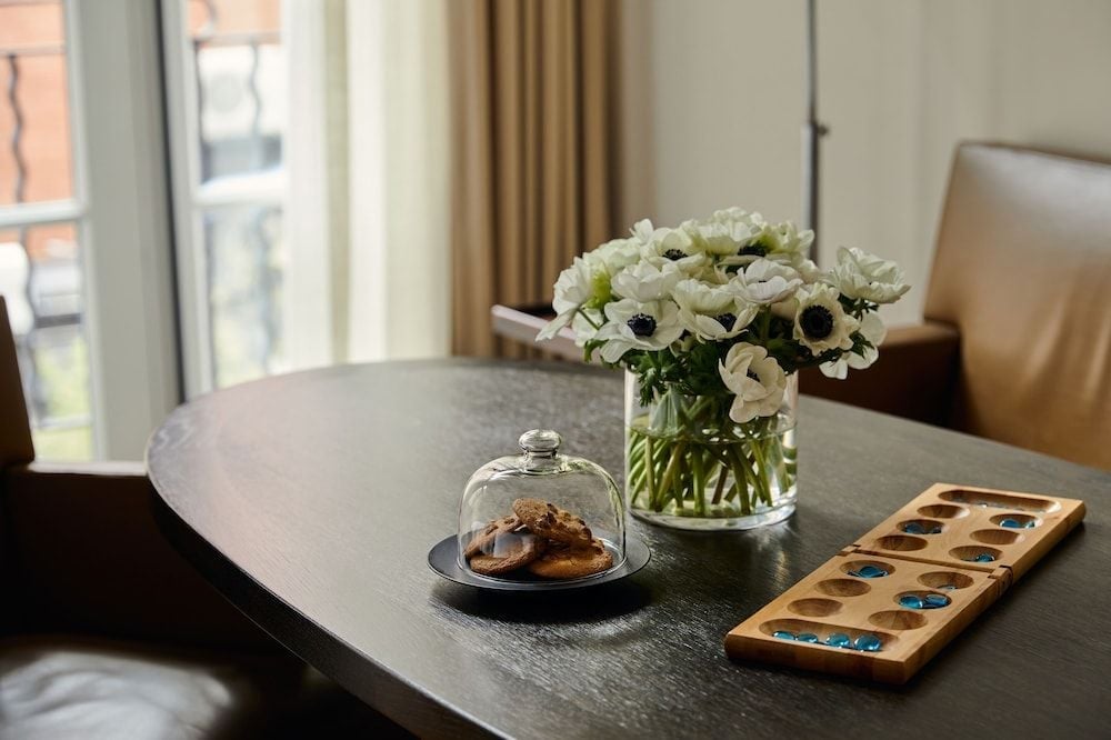 White anemones with dark centers grace a sleek oval table alongside artisanal treats under glass and a handcrafted mancala board, creating intimate in-room luxury.