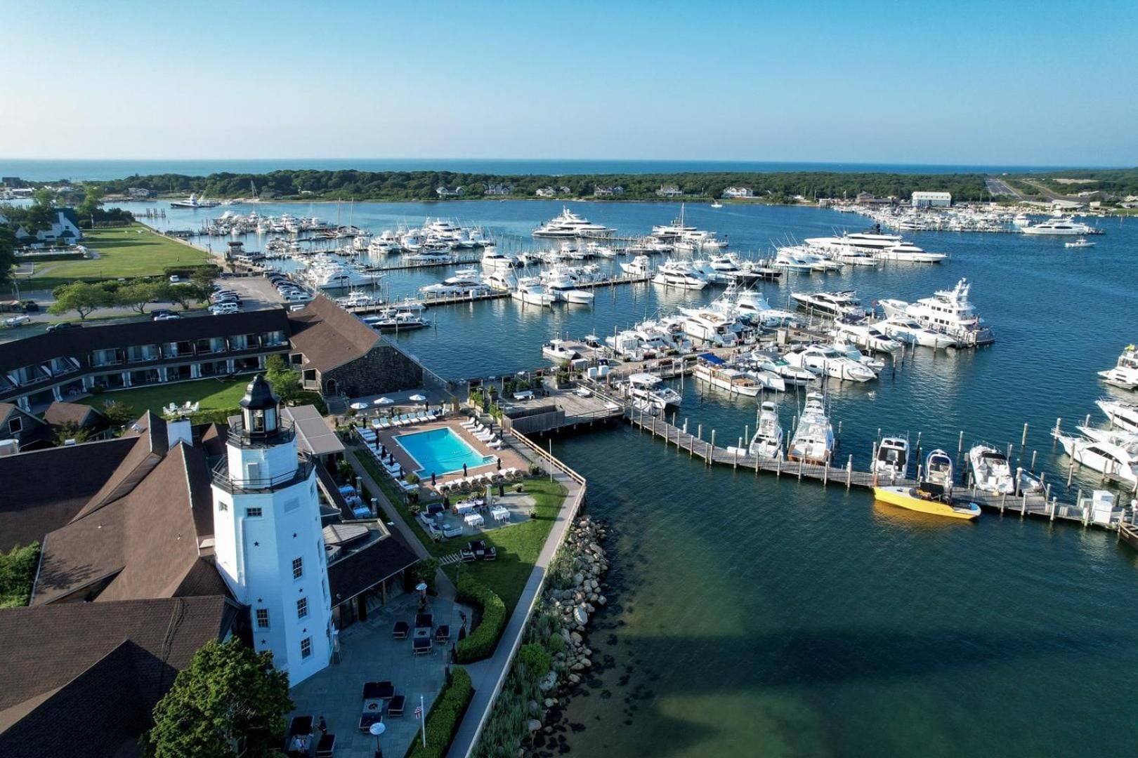 Historic white lighthouse stands sentinel over crystalline waters where luxury yachts dock beside inviting poolside loungers, creating an idyllic maritime escape on Long Island's eastern tip.