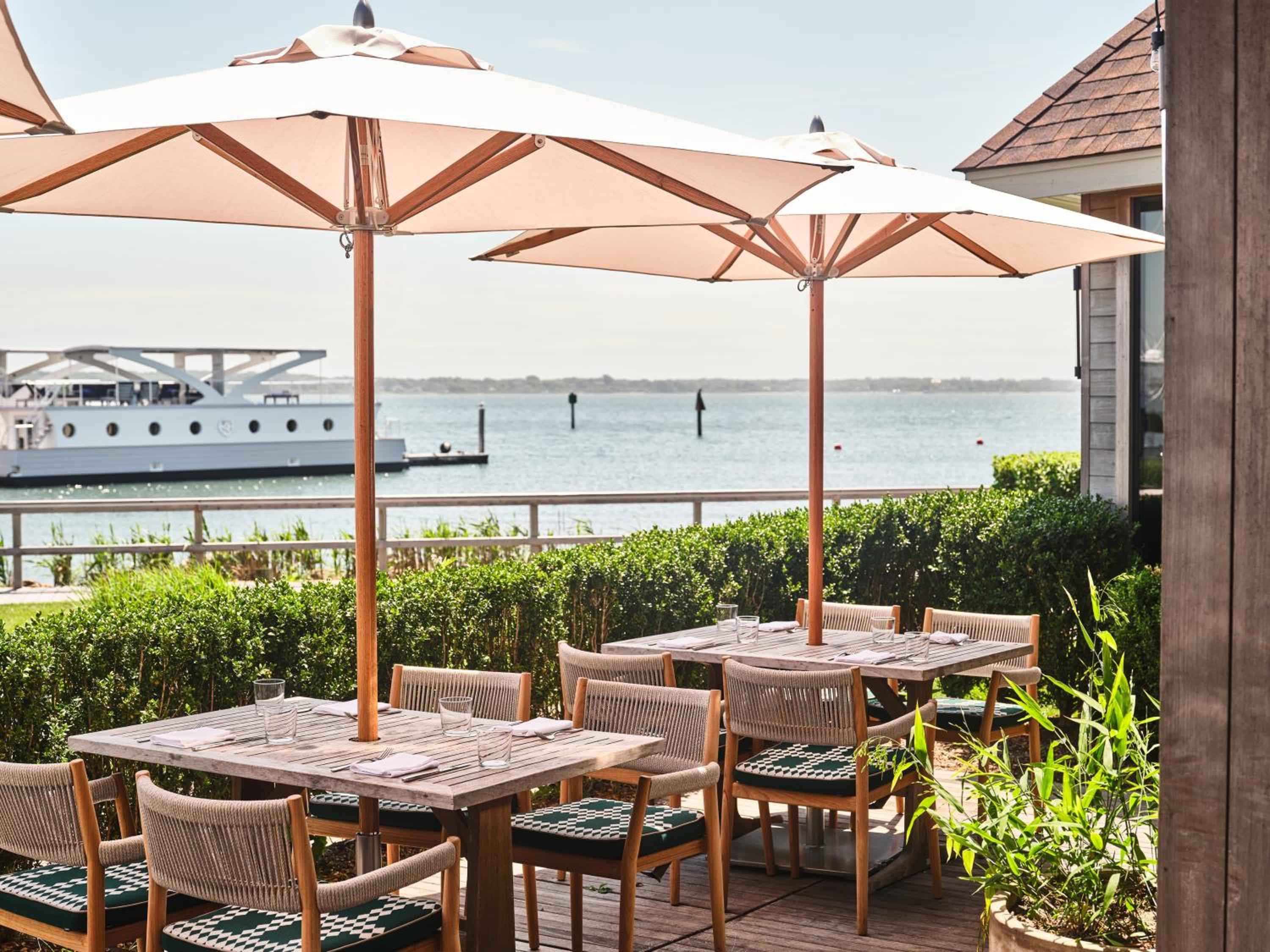 Waterfront dining unfolds beneath cream umbrellas, where teak tables overlook yacht-dotted waters and salt air mingles with the promise of exceptional cuisine.