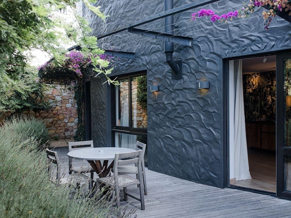 Sculptural charcoal walls create dramatic texture against purple bougainvillea, while intimate dining unfolds on weathered decking overlooking terraced stone gardens.