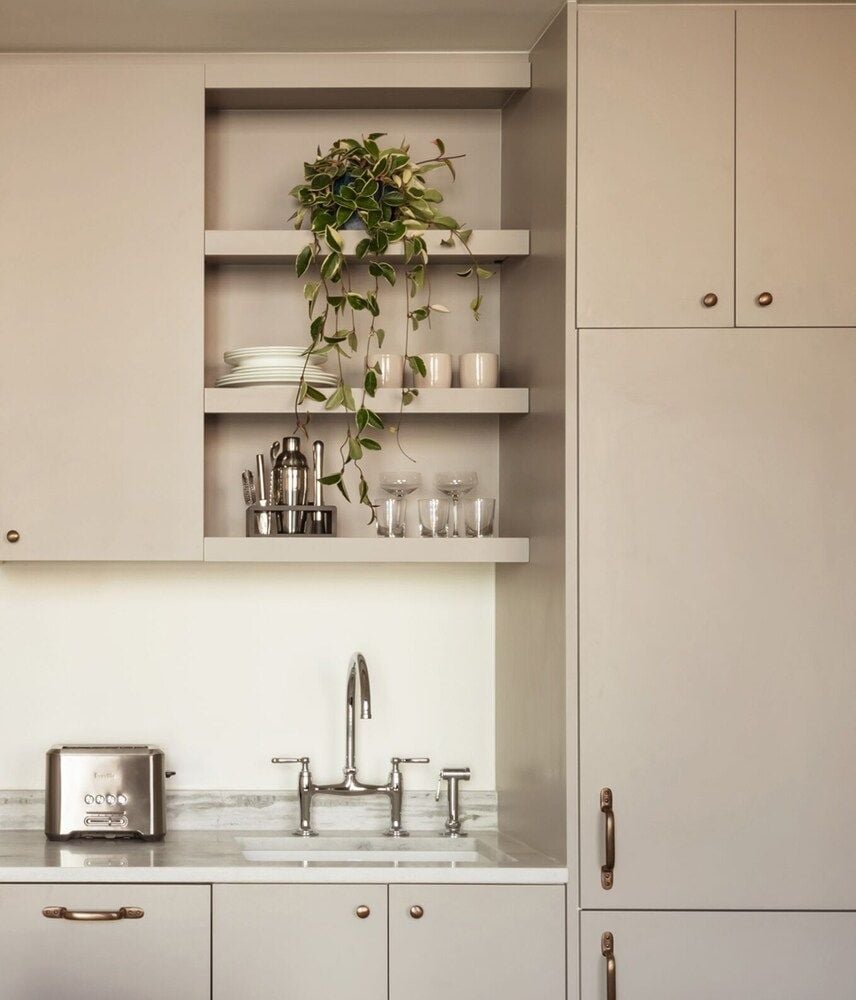 Creamy cabinetry and brass fixtures create a serene kitchen vignette, where trailing greenery softens clean lines and vintage-inspired hardware adds warmth to minimalist design.