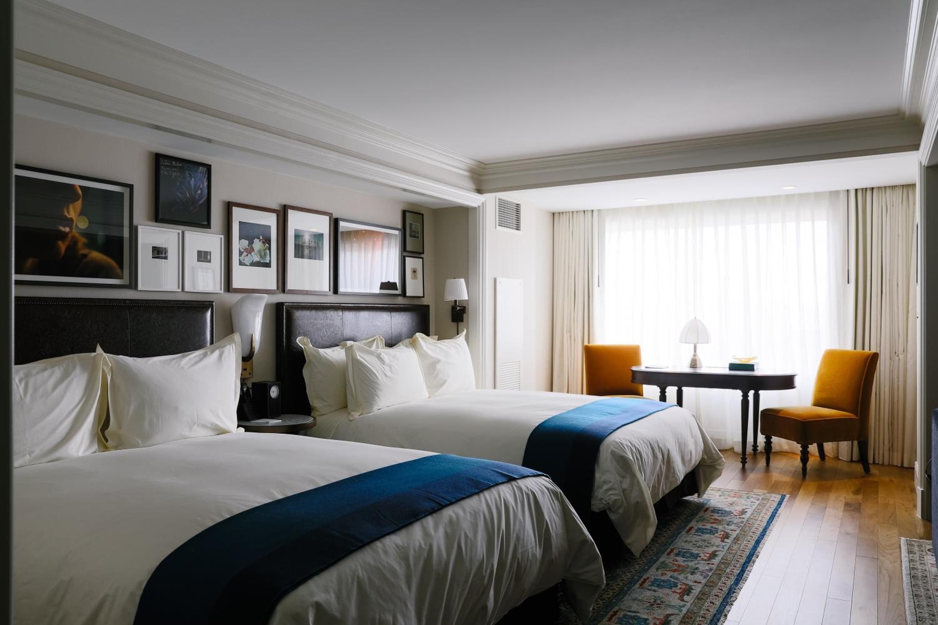 Twin beds dressed in crisp white linens and navy runners create an intimate sanctuary, while gallery walls and golden accent chairs add sophisticated warmth to this thoughtfully appointed guest room.