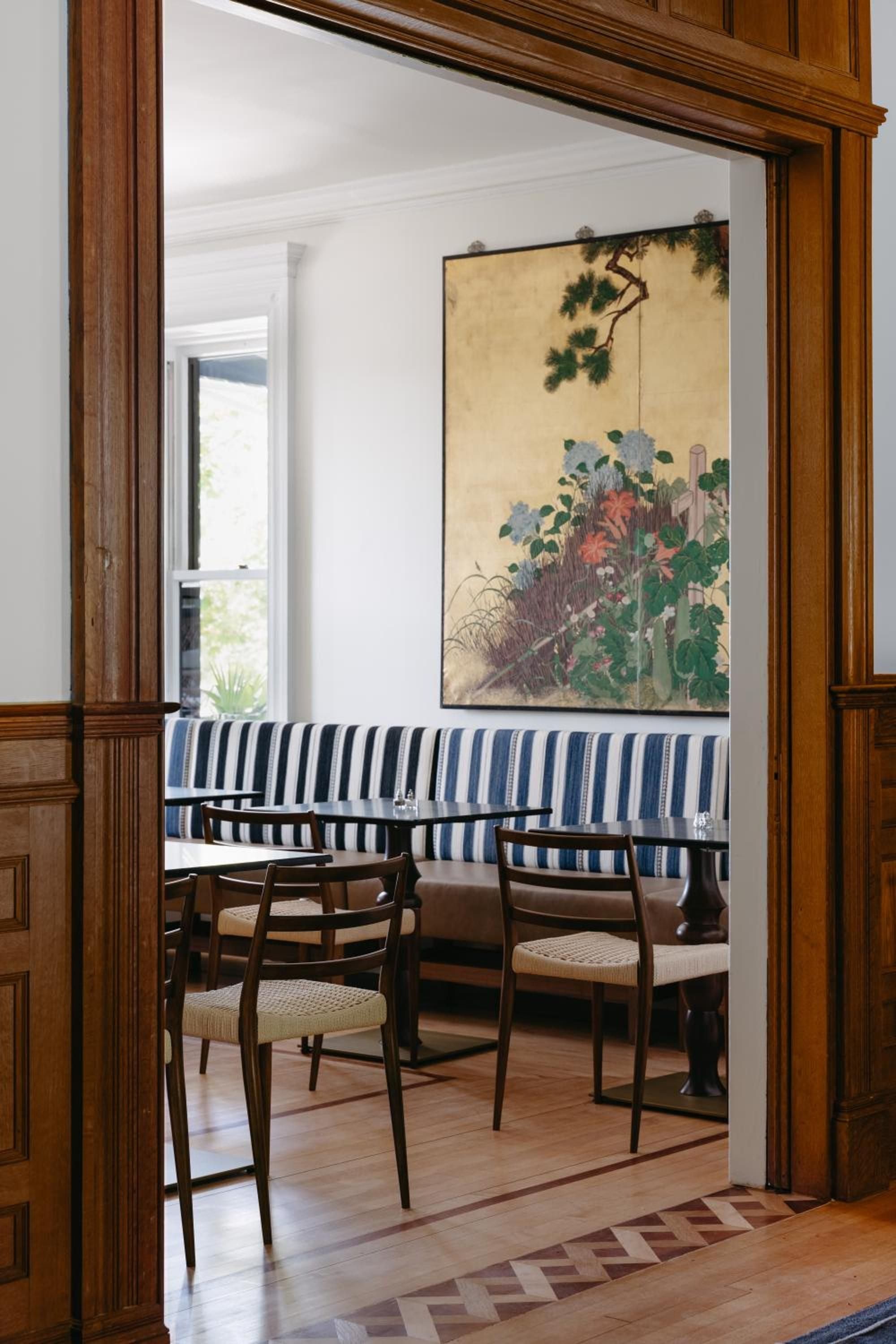 Nautical-striped banquettes and vintage botanical artwork create an intimate dining alcove, where warm wood frames and woven chair seats invite leisurely coastal conversations.