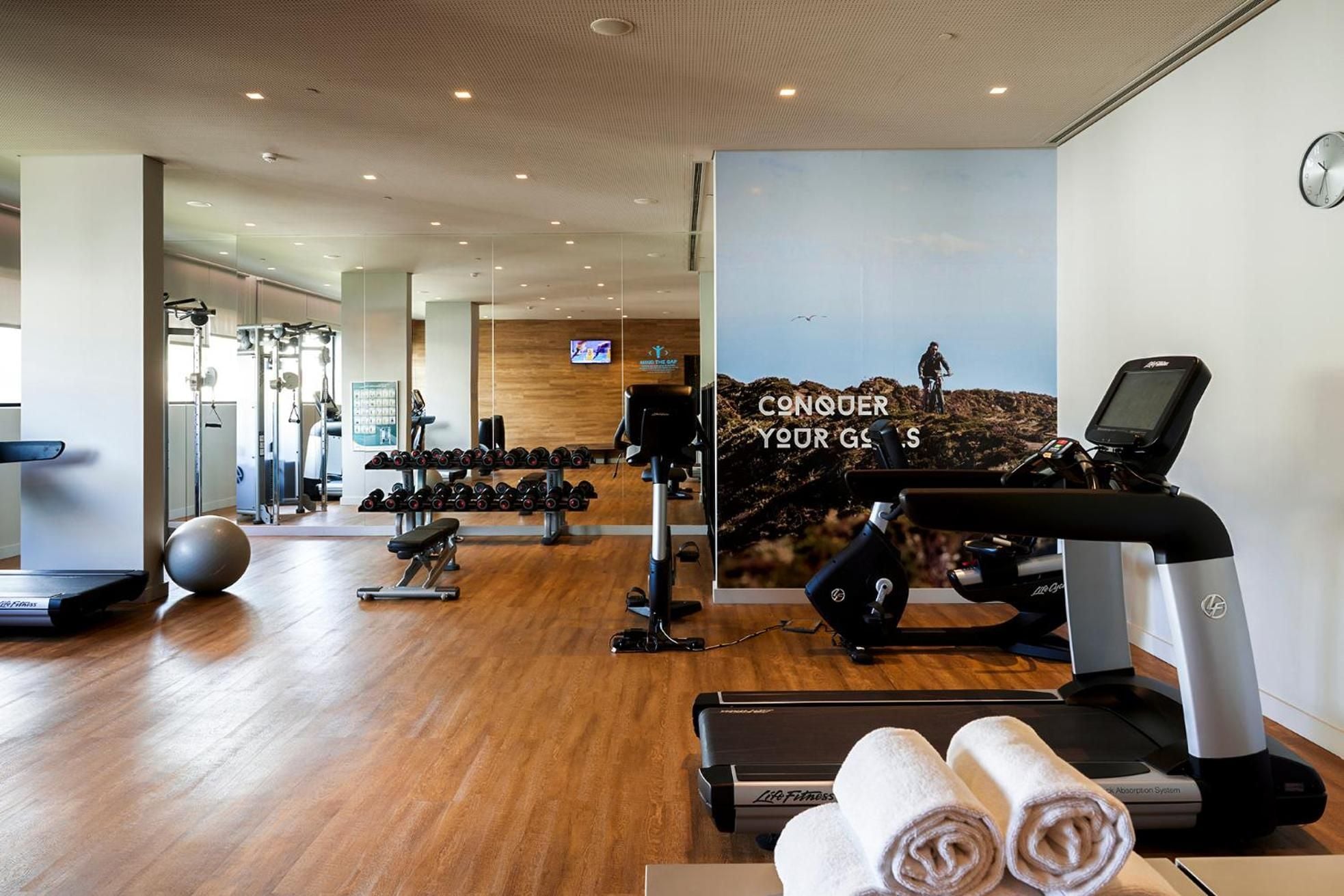 Modern cardio equipment faces an inspiring mountain biking mural, while polished wood floors and crisp white towels create an upscale athletic sanctuary with motivational edge.