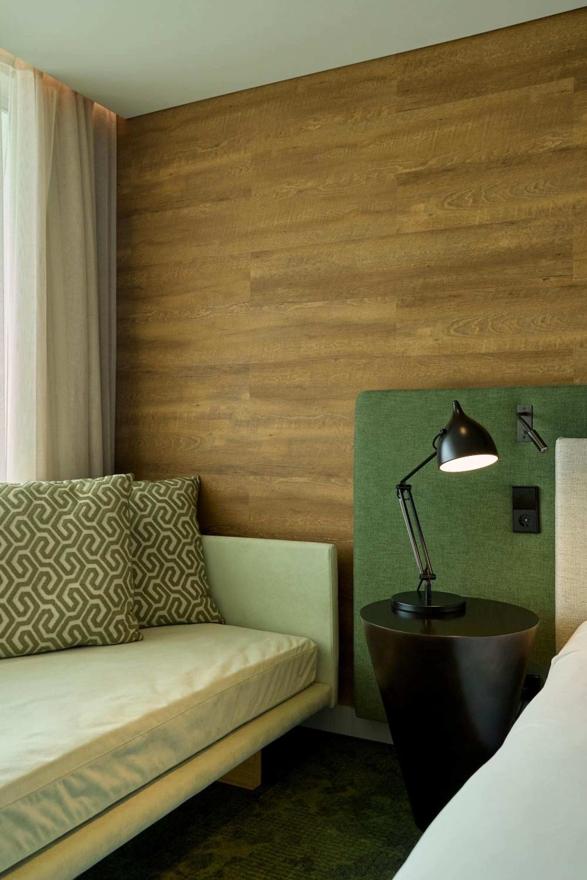 Warm wood paneling meets sage green accents in thoughtfully designed guest quarters, where geometric pillows and sleek task lighting create an inviting reading nook.