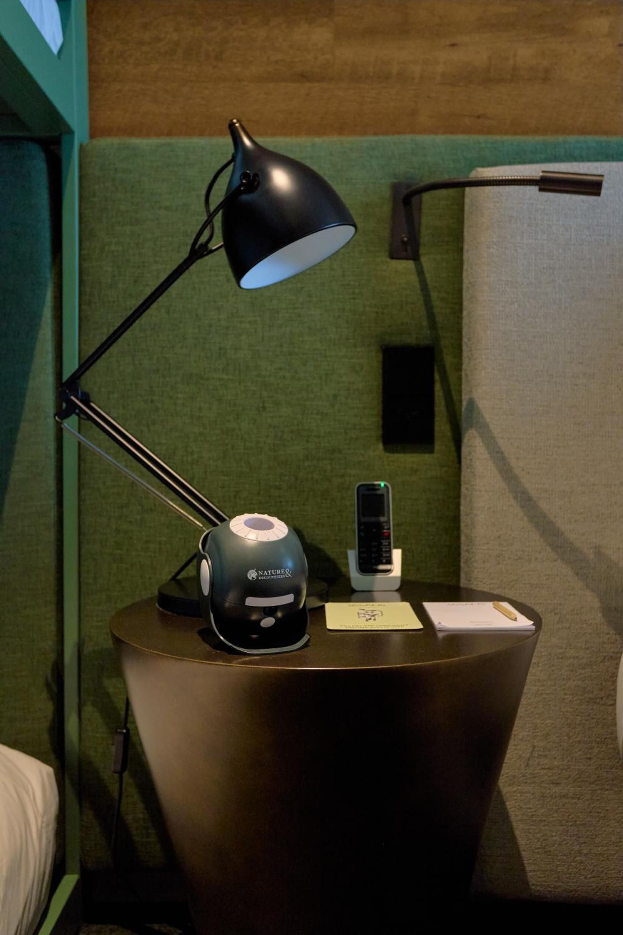 Thoughtful bedside essentials include a sleek adjustable lamp, cordless phone, and coffee service on warm wood surfaces against sage-green accent walls.