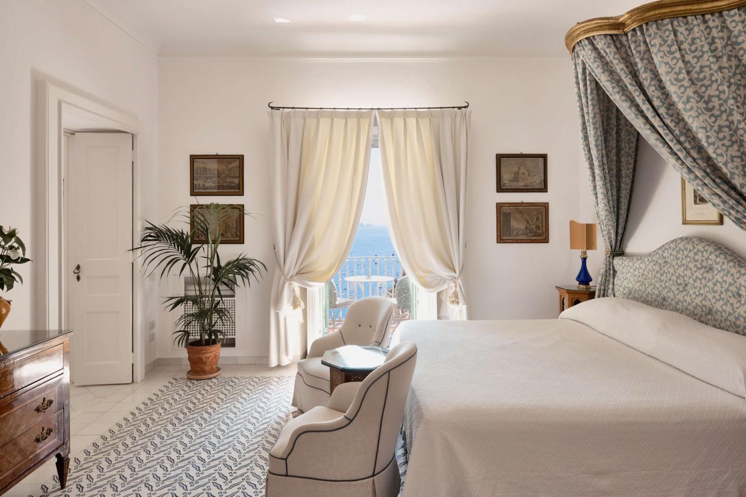Ornate canopy bed frames azure Amalfi Coast views while antique prints and patterned fabrics create an intimate sanctuary of old-world Mediterranean elegance.