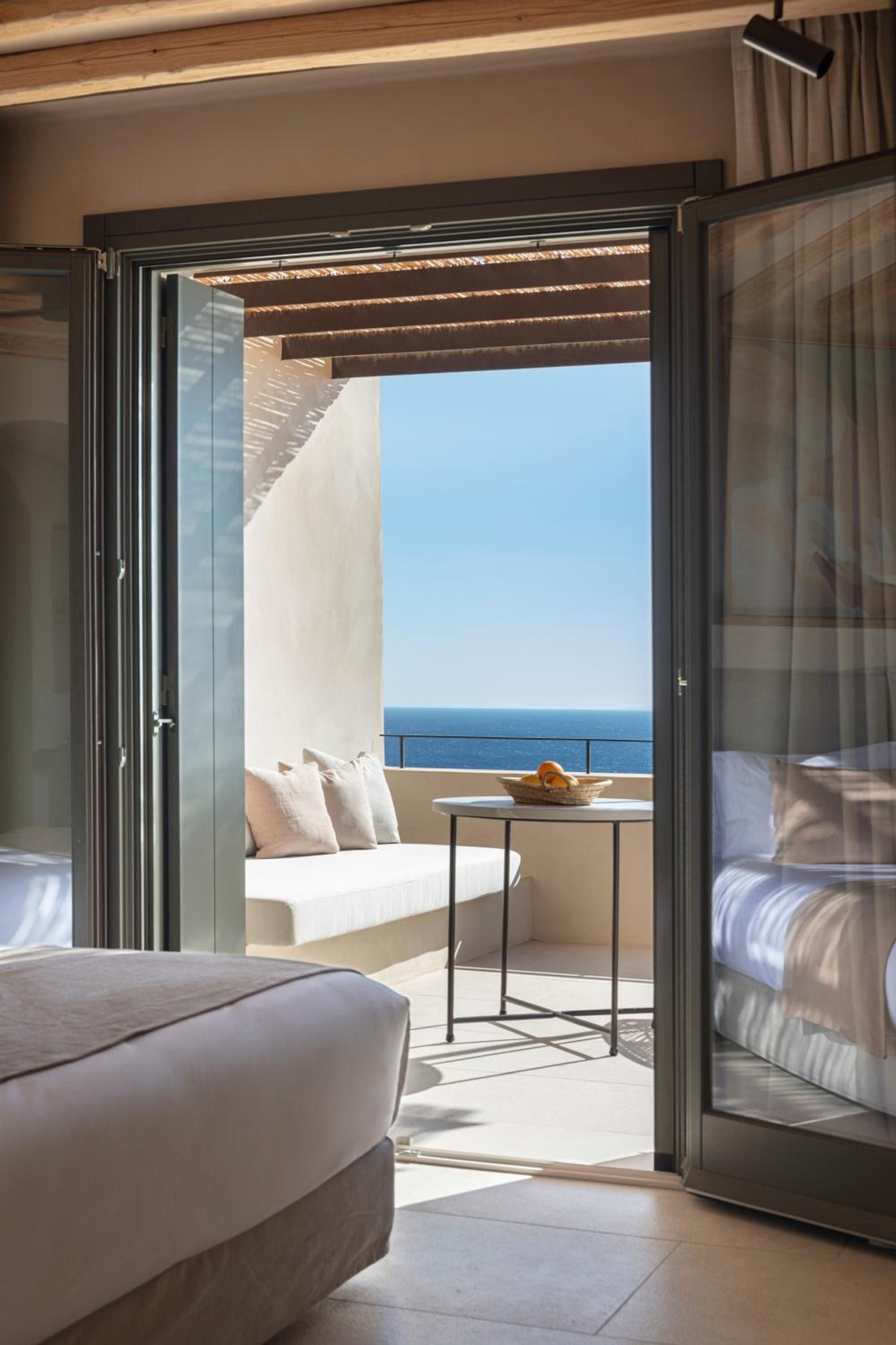 Natural light floods the bedroom through floor-to-ceiling glass doors, revealing a private terrace where whitewashed stone meets endless Aegean blue.