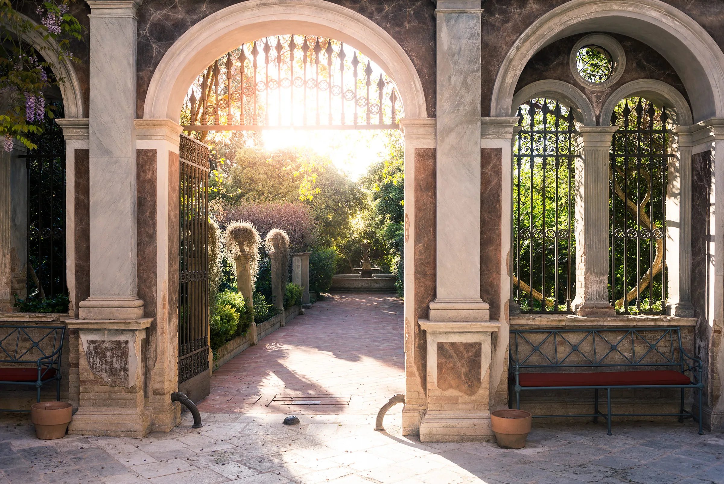 Stone archways frame sunlit pathways through Palazzo Margherita's classical courtyard, where weathered columns and wrought-iron details create an enchanting passage into this Basilicata retreat.