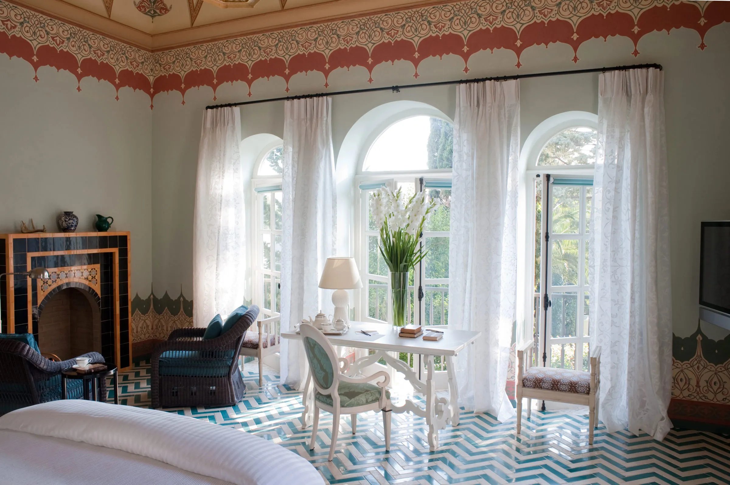 Ornate Moroccan-inspired frescoes crown a luminous suite where arched French doors frame verdant gardens, while geometric tiles and antique furnishings create an enchanting retreat.