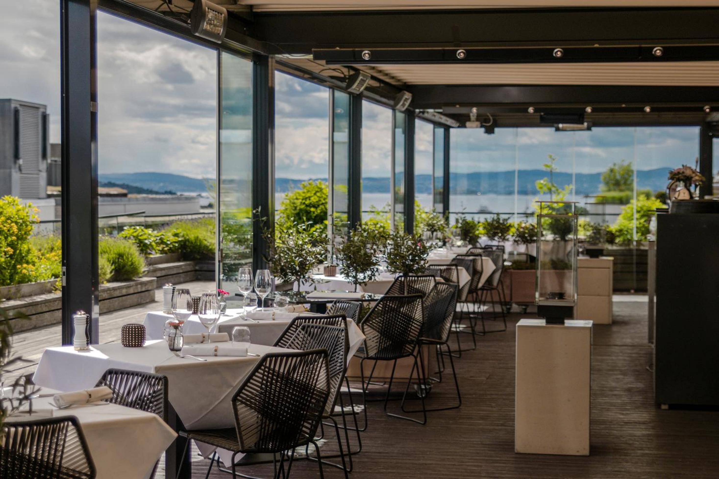 Floor-to-ceiling windows frame Oslo's fjord while diners settle into sleek wicker chairs beneath string lights, creating an intimate terrace dining experience above the city.