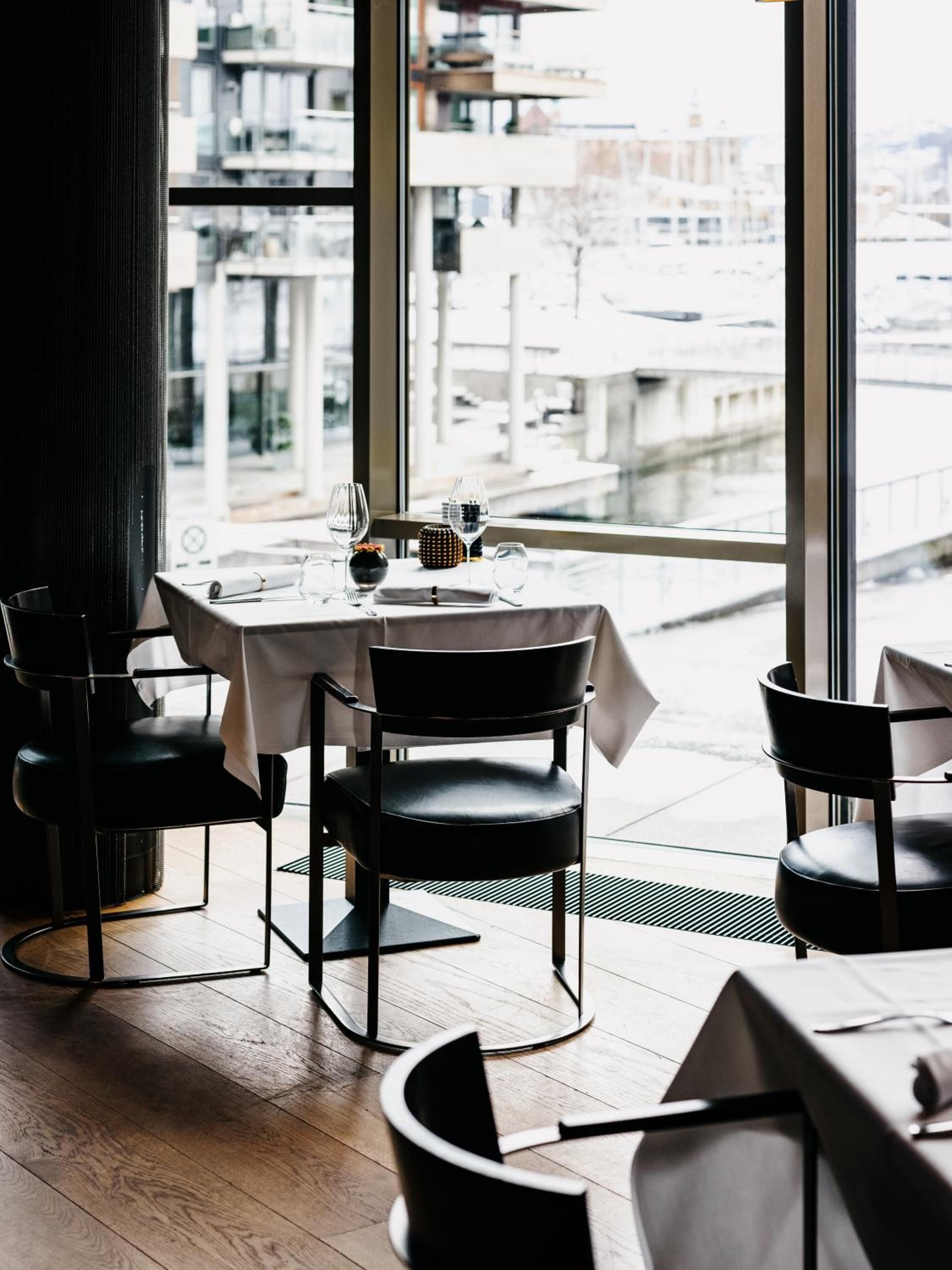 Floor-to-ceiling windows frame Oslo's harbor while crisp linens and sculptural black chairs create an intimate dining sanctuary where Nordic minimalism meets waterfront sophistication.