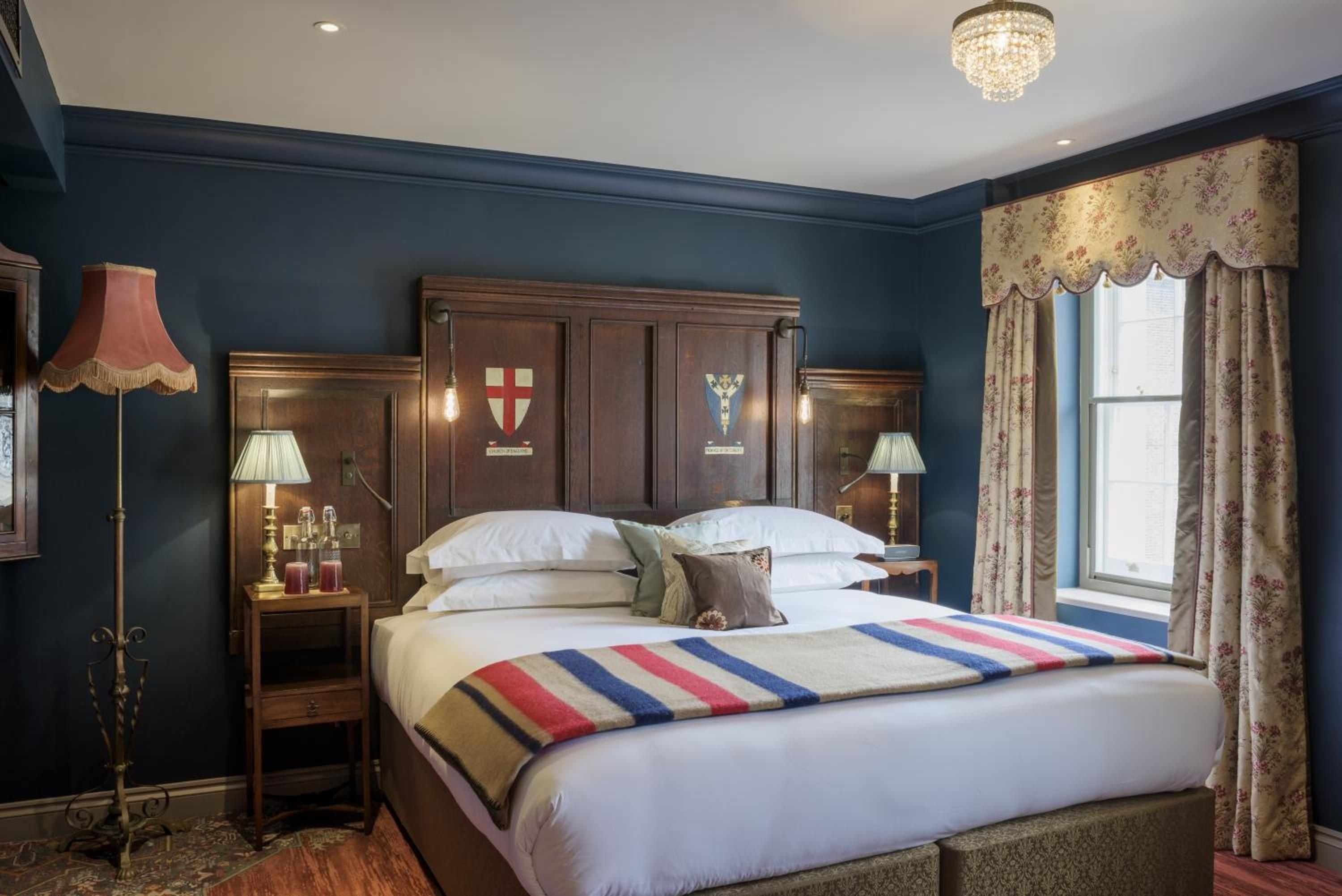 Navy walls embrace a mahogany headboard adorned with heraldic crests, while crystal chandeliers cast golden light across striped throws and floral curtains in this quintessentially British retreat.