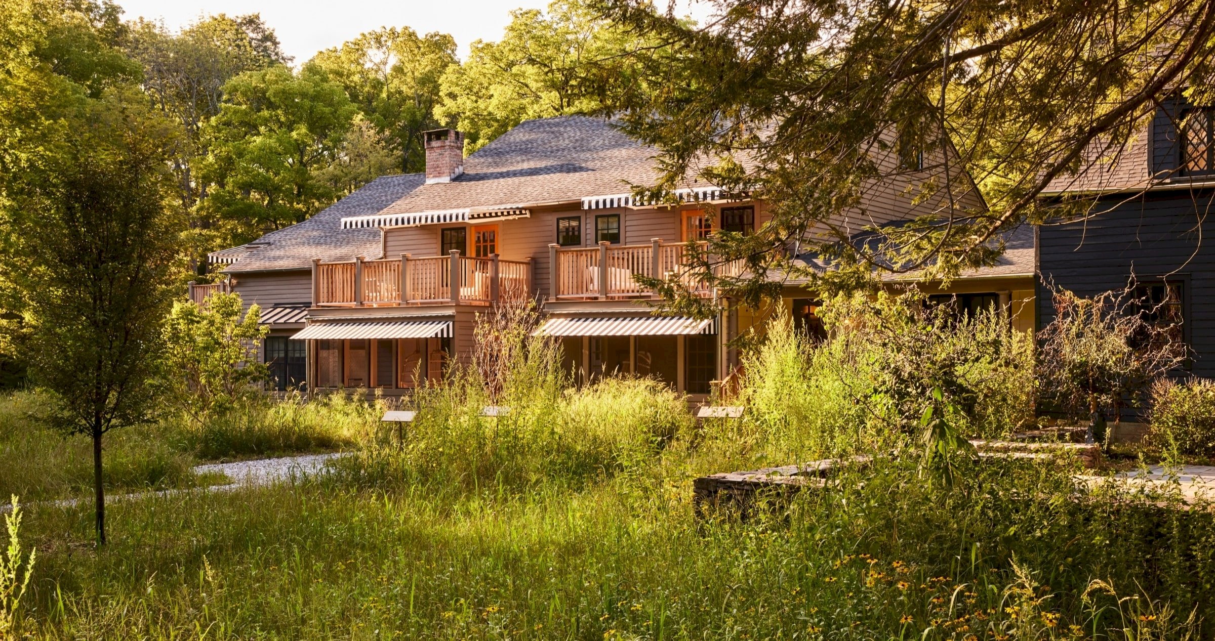 Weathered cedar shingles and striped awnings peek through wild meadow grasses, where Troutbeck's main house settles into the Berkshire landscape like a beloved summer memory.