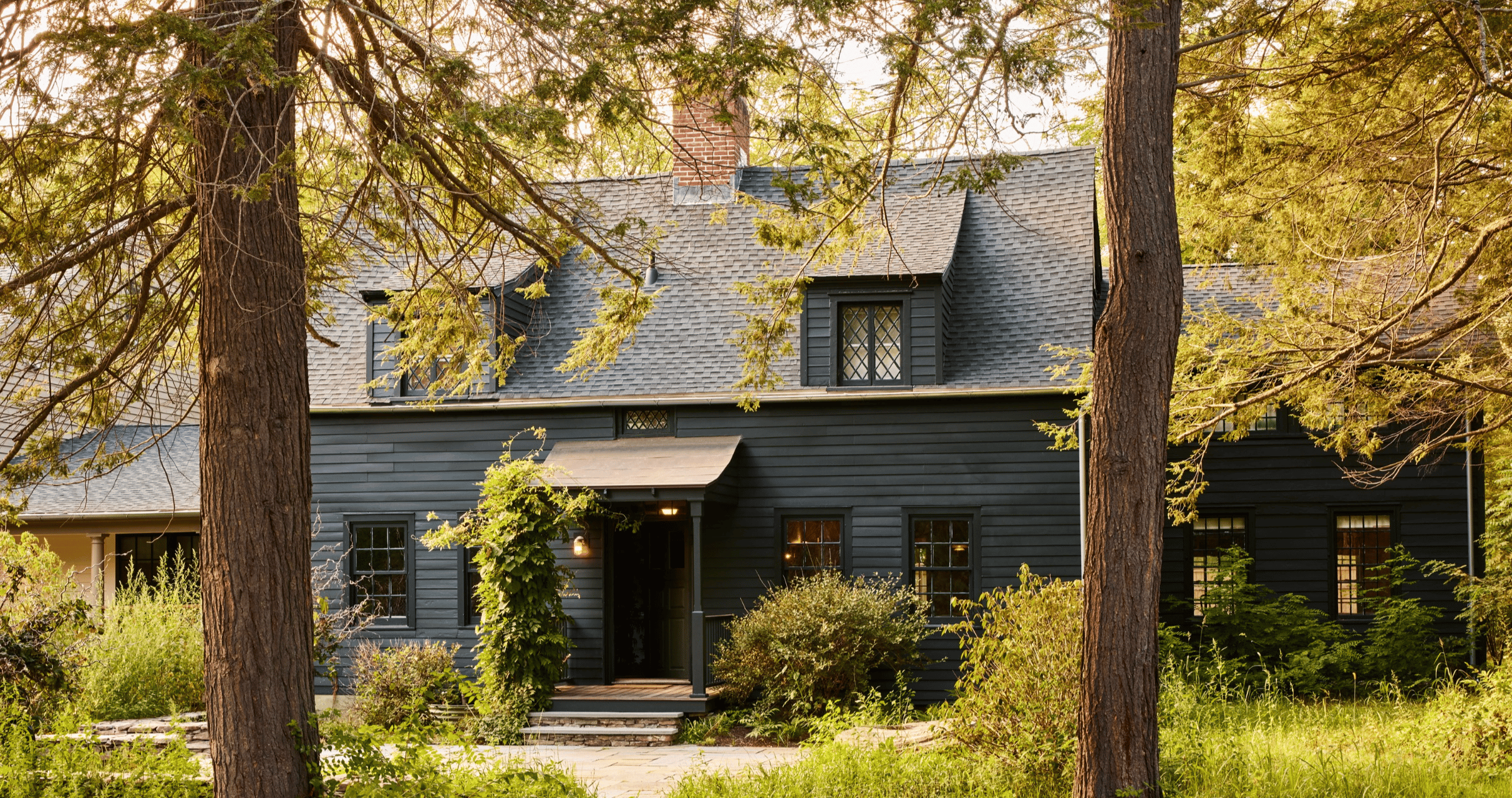Weathered cedar shingles and diamond-pane windows peek through ancient maples, where dappled golden light creates an enchanting approach to this historic Berkshires retreat.
