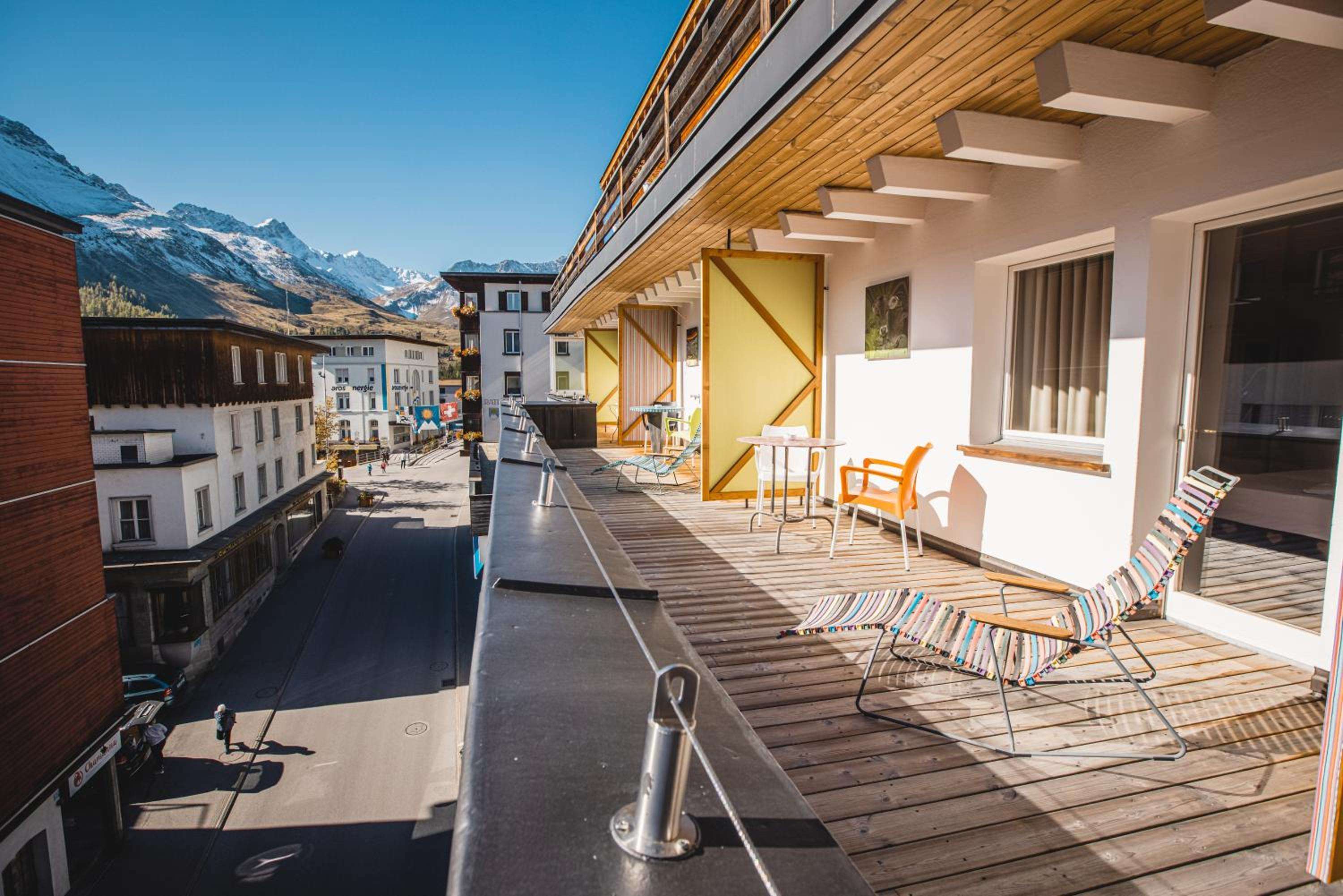Colorfully striped lounge chairs dot the wooden terrace, where Alpine peaks frame this sun-soaked retreat high above the village streets below.