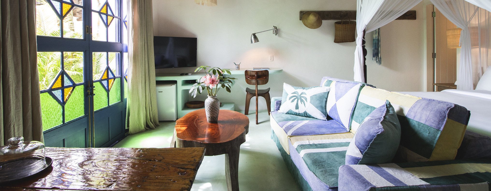 Sunlight streams through geometric stained glass onto a tranquil suite where tropical colors meet rustic wooden furnishings and flowing white mosquito netting.