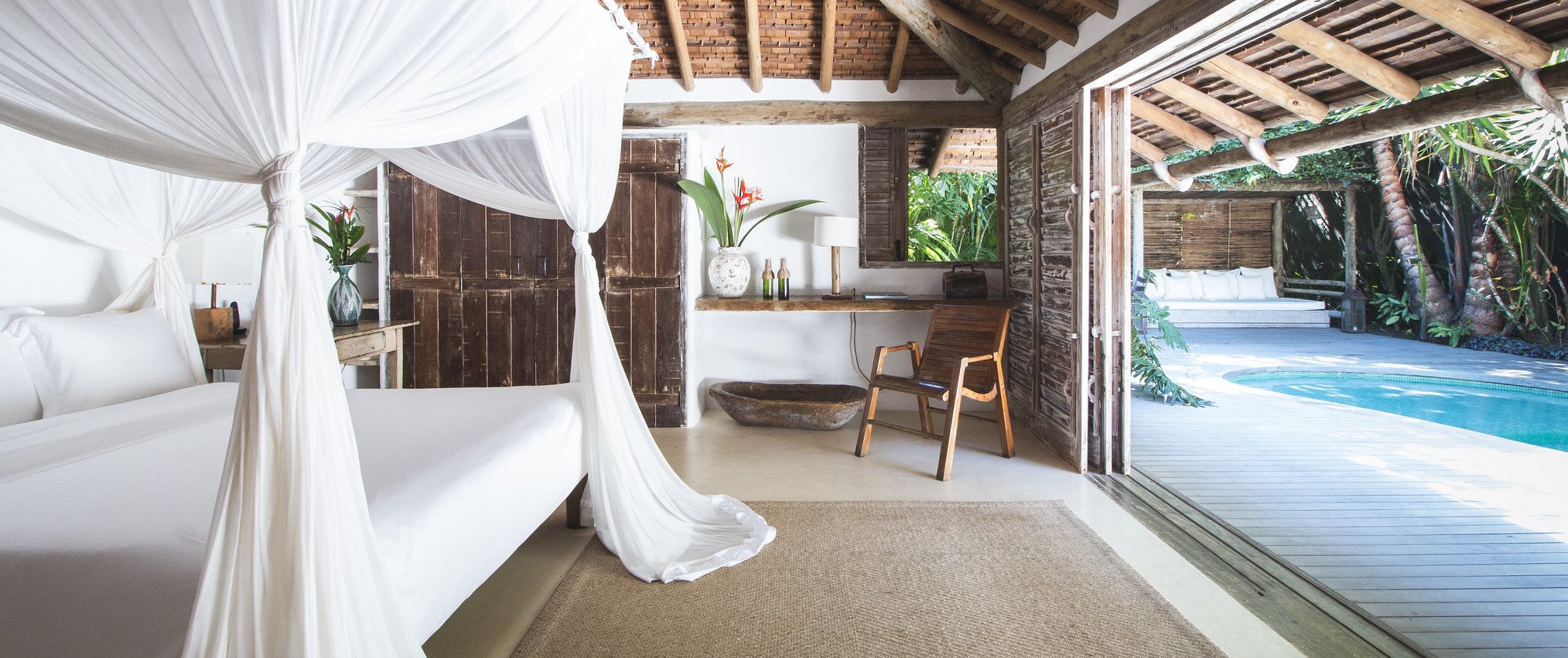 Flowing white curtains frame a canopy bed in this open-air suite, where rustic wood beams meet polished concrete and tropical breezes drift in from the azure pool beyond.