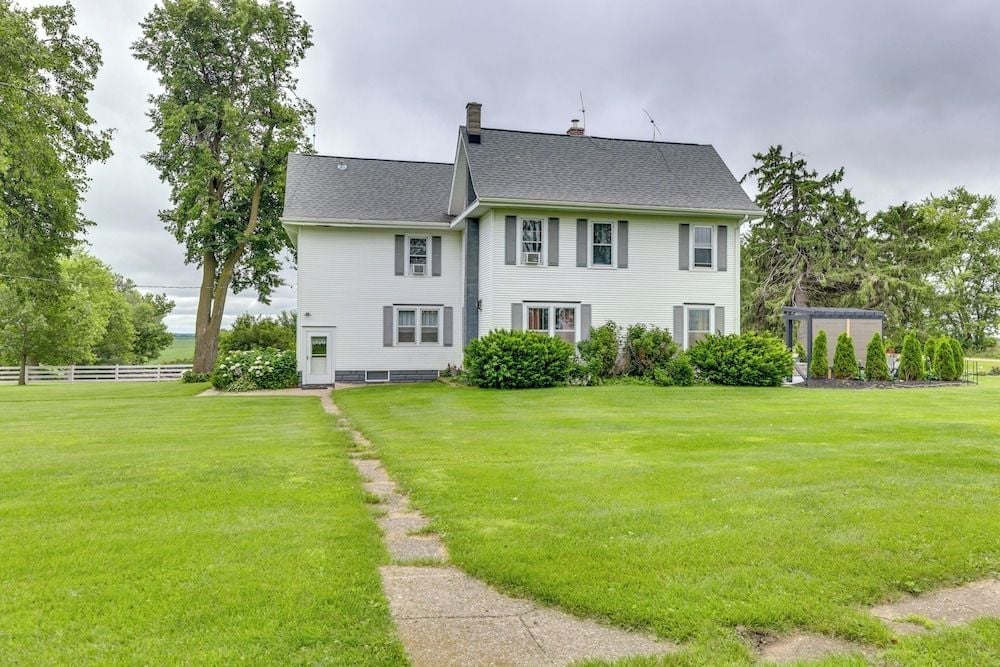 Pristine lawns stretch toward this charming white colonial farmhouse, where gray shutters frame windows and mature trees create a pastoral sanctuary under moody skies.