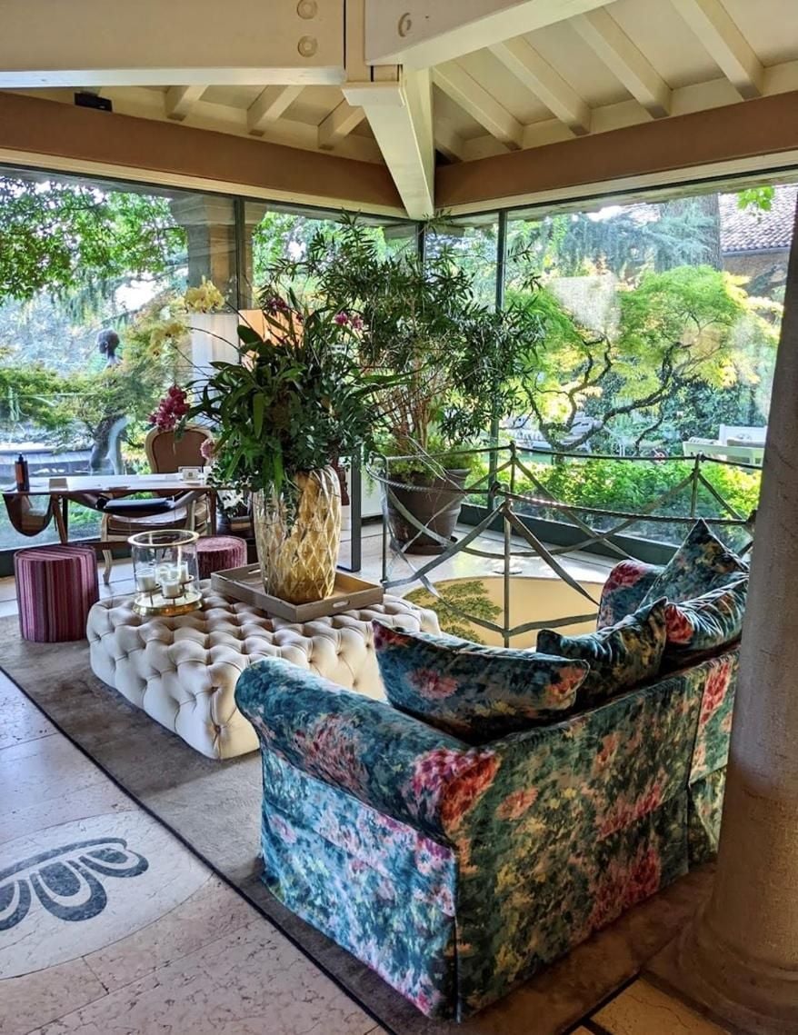 Vibrant floral armchairs and tufted ottomans create an inviting conversation corner beneath exposed beams, while floor-to-ceiling windows frame verdant hillside gardens beyond.