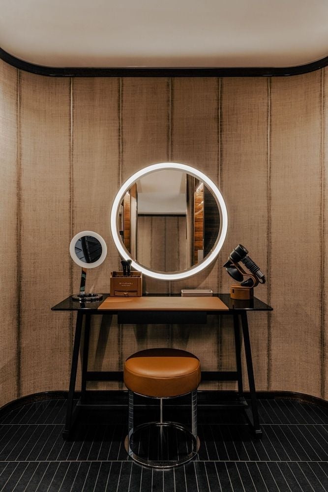 Illuminated vanity mirrors cast a warm glow against textured bamboo panels, while brass accents and leather seating create an intimate grooming sanctuary.