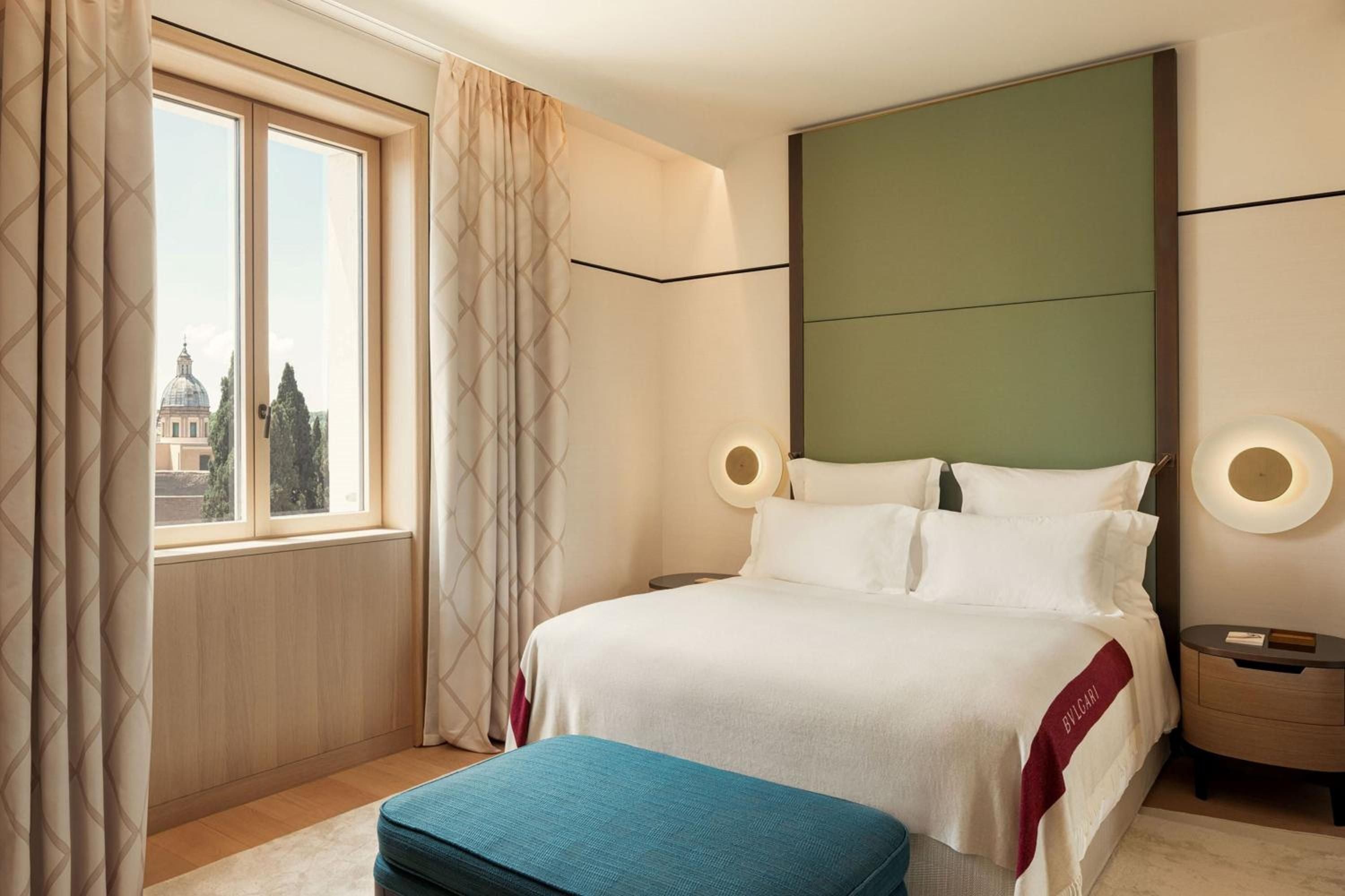 Sage-green headboards and warm travertine walls create serene sanctuaries overlooking Rome's historic rooftops, where circular bedside sconces cast gentle pools of light.
