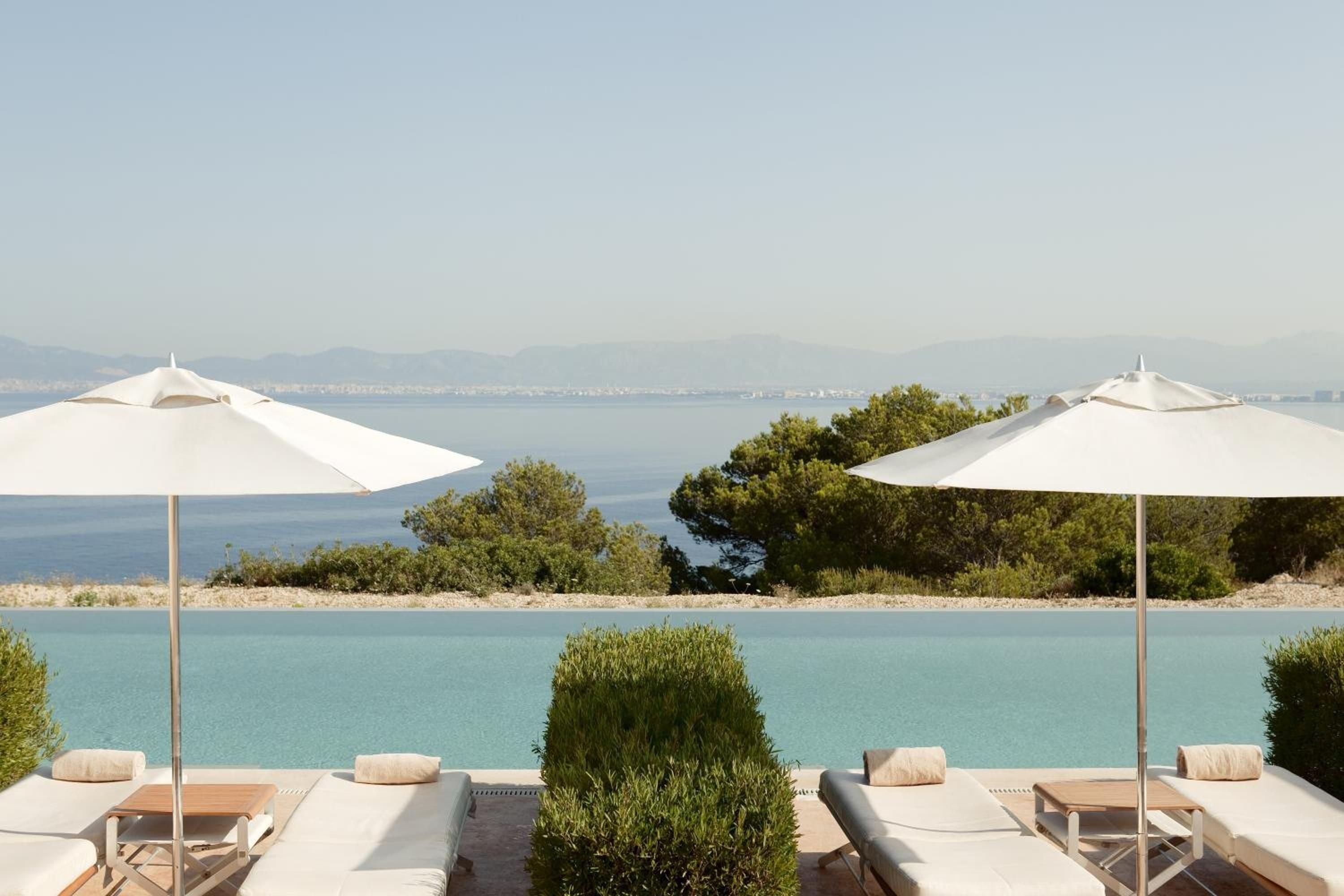 White umbrellas cast crisp shadows over infinity pool loungers, where manicured hedges frame turquoise waters that seem to spill into the Mediterranean beyond.