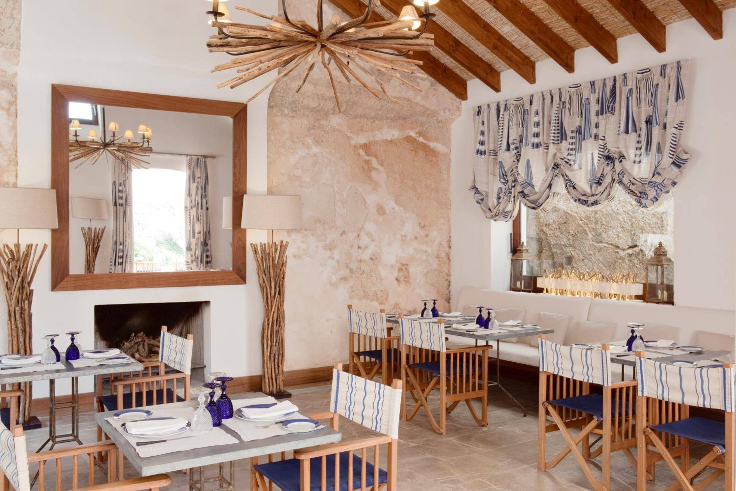 Sunlight filters through blue-striped curtains onto rustic wooden tables in Cap Rocat's intimate dining room, where exposed stone walls and driftwood chandeliers create Mediterranean warmth.