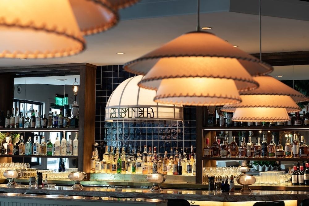 Warm pendant lights cast a golden glow over Casa Duro's sophisticated bar, where polished shelves display an impressive collection of spirits behind gleaming copper stools.