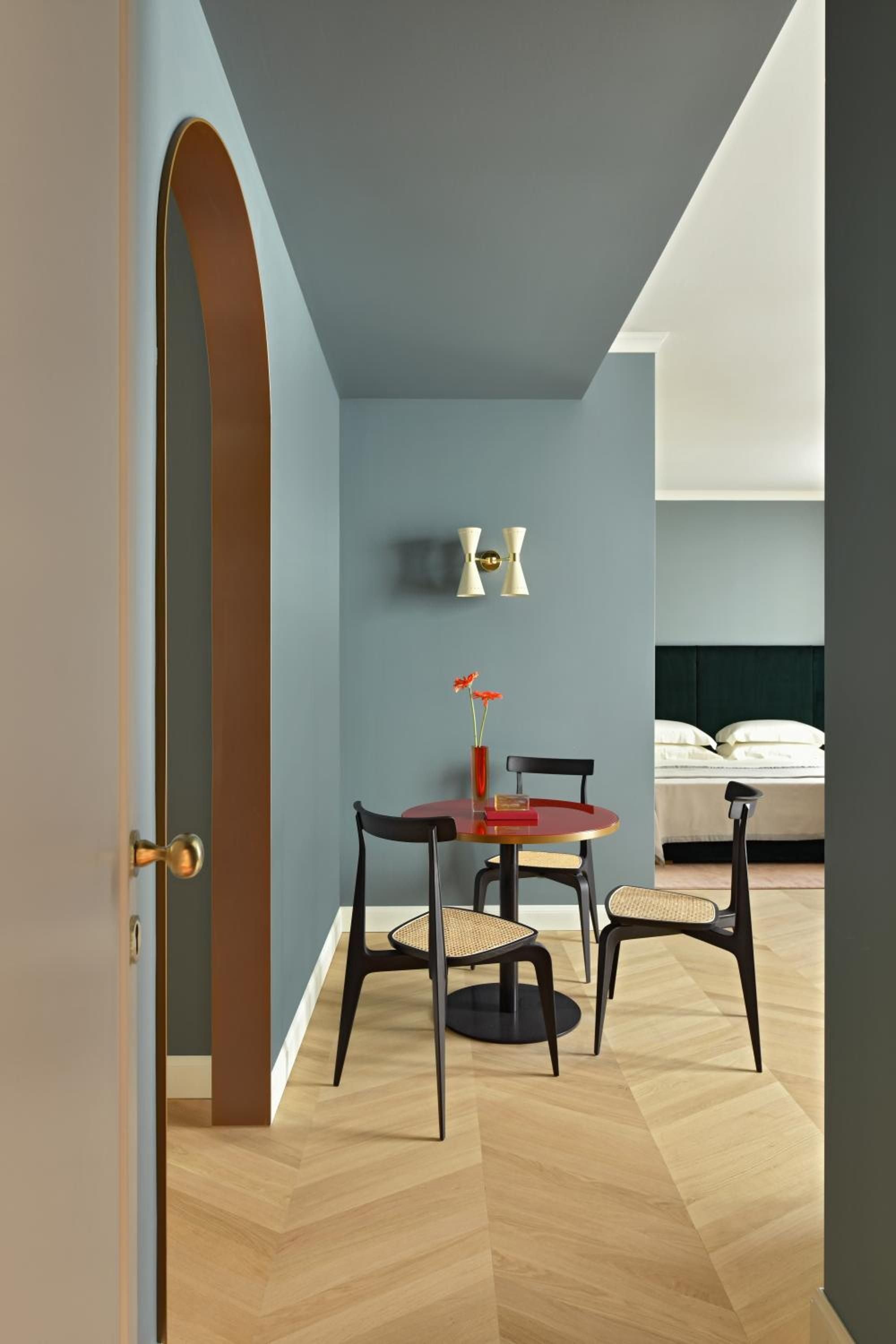 Soft blue walls frame a cozy dining nook where mid-century chairs gather around a crimson table, while warm parquet flows beneath this intimate corner retreat.