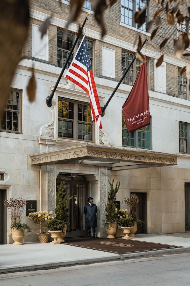 A uniformed doorman stands beneath ornate limestone details and burgundy flags, where Fifth Avenue sophistication meets old-world hospitality at this Upper East Side gem.