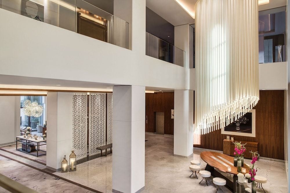 Soaring double-height ceilings frame a dramatic cascading light installation, while warm walnut accents and geometric screens create intimate conversation pockets below the mezzanine level.