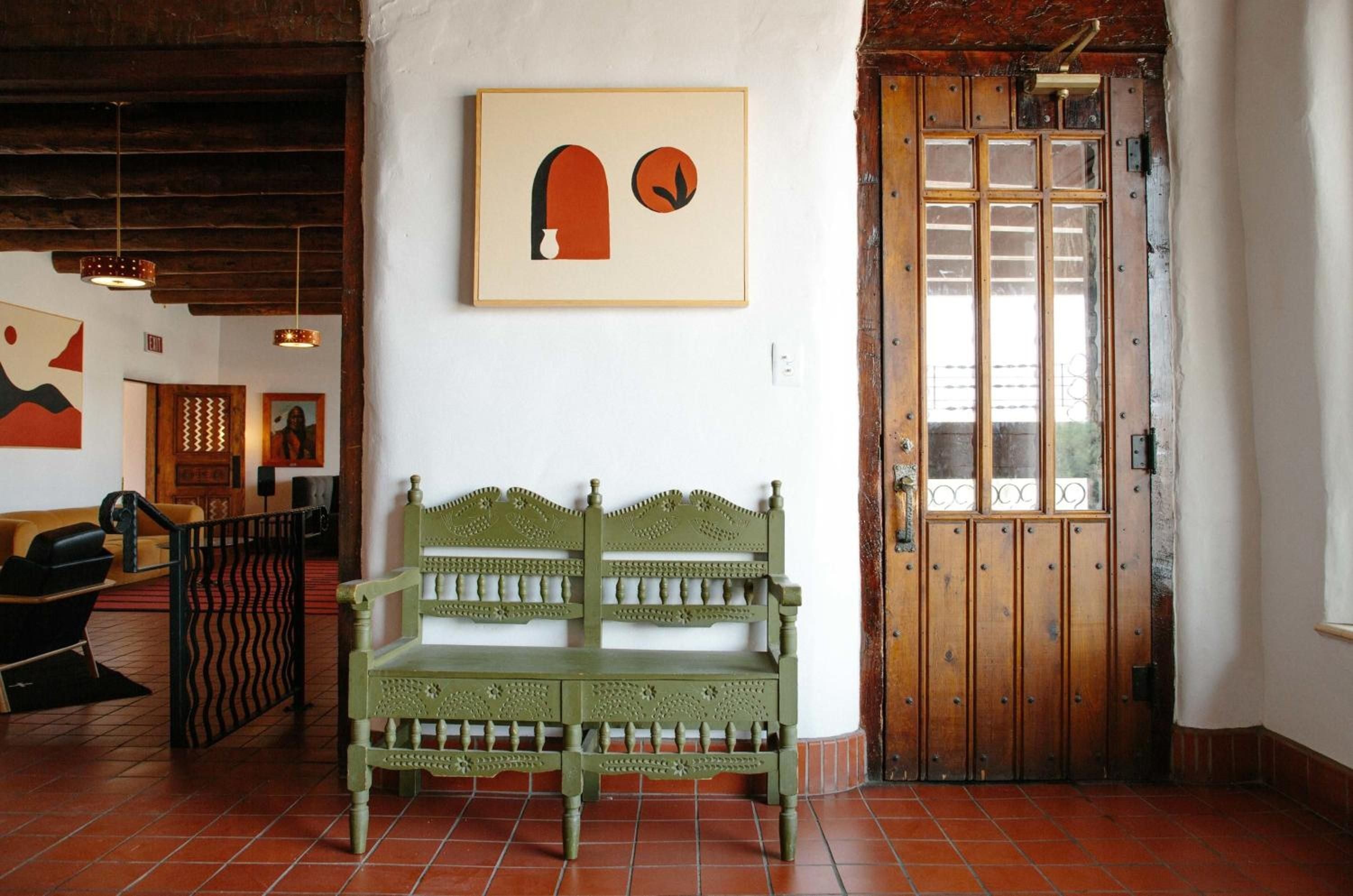 Hand-carved green wooden chairs anchor a sun-drenched corner where terra cotta tiles meet whitewashed walls adorned with minimalist geometric art and rustic wooden shutters.