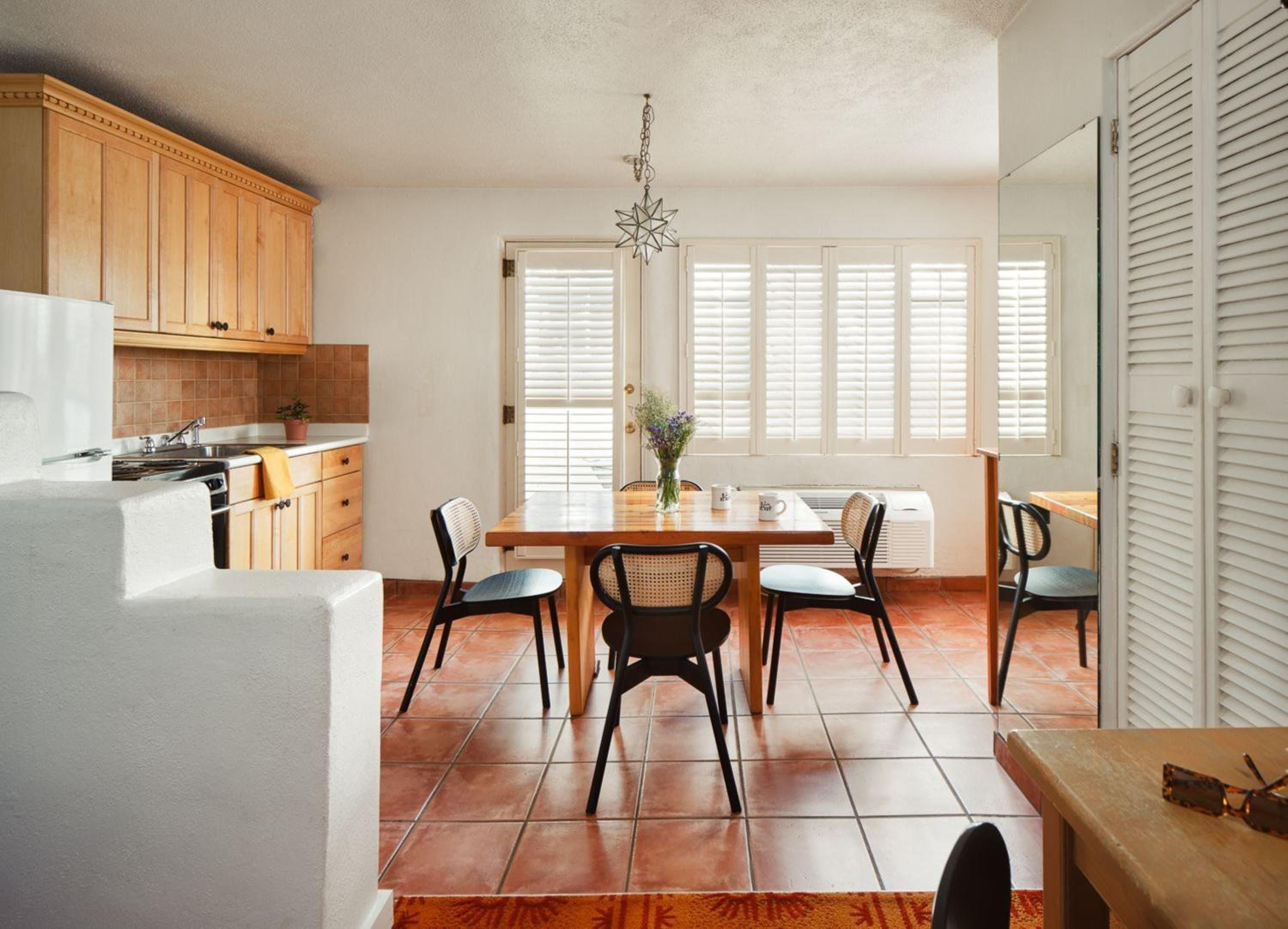 Terracotta tiles warm the open kitchen and dining area, where mid-century chairs gather around a honey-toned table beneath desert light filtering through plantation shutters.