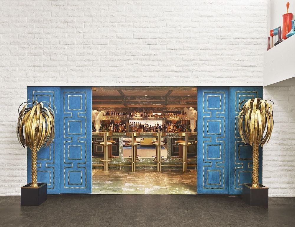 Bold cobalt doors adorned with golden studs open into the hotel's glamorous dining scene, where towering palm-frond sculptures frame a sophisticated bar awash in warm amber light.