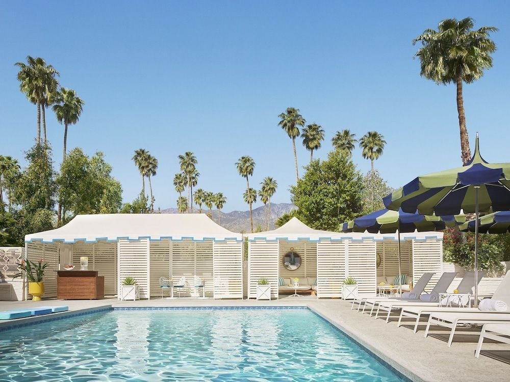 Mid-century modern poolside charm meets desert glamour with striped umbrellas, white lounge chairs, and towering palms framing the San Jacinto Mountains beyond.