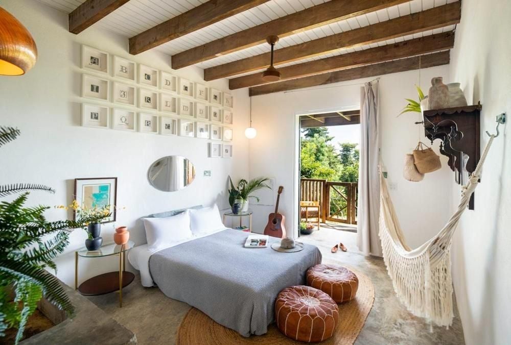 Exposed wooden beams frame this serene bedroom where macramé hammocks and leather poufs create bohemian charm against crisp white linens and jungle views.