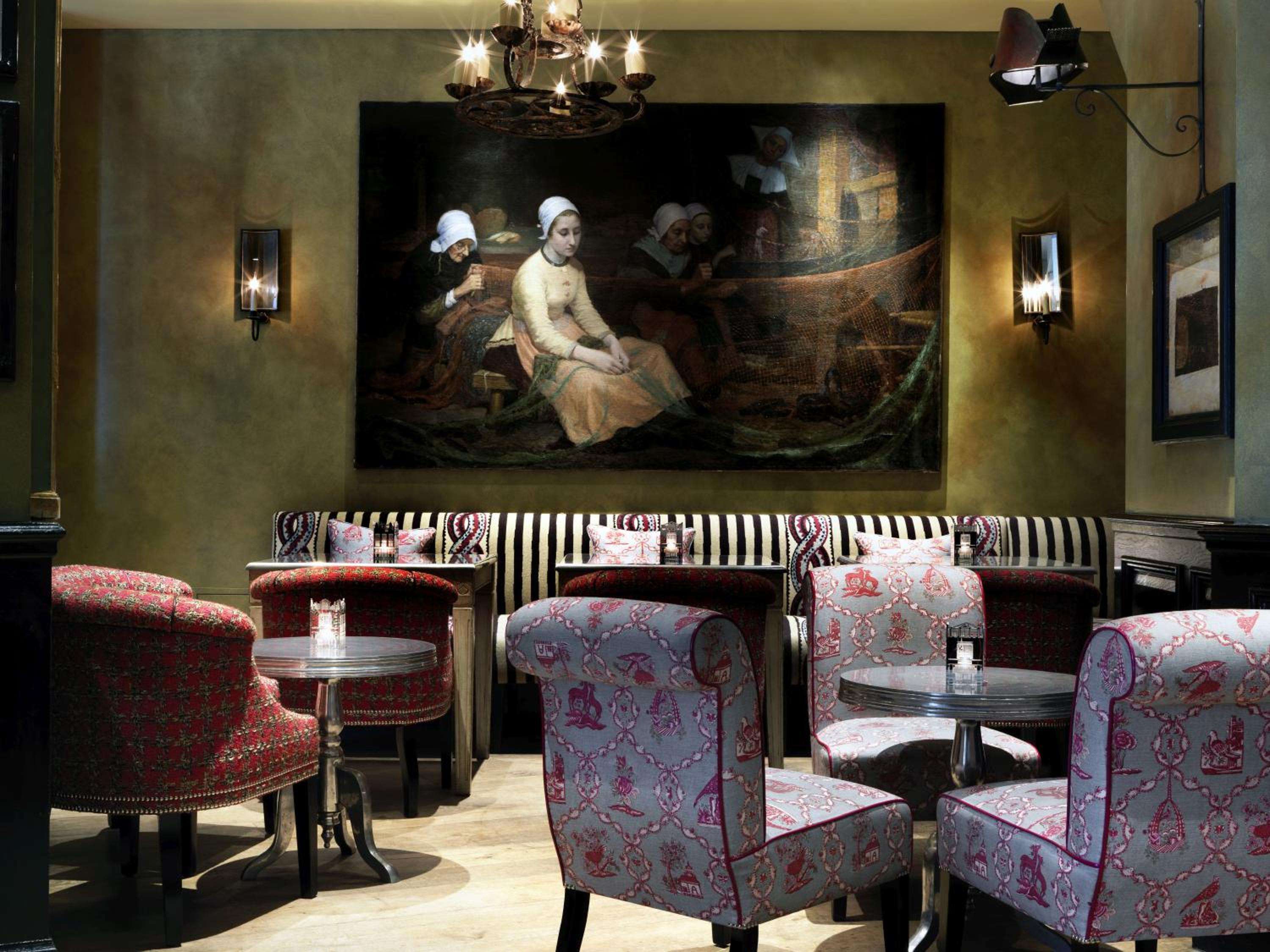 Jewel-toned armchairs and dramatic maritime paintings create an intimate drinking den where classical elegance meets theatrical London flair beneath flickering candlelit chandeliers.
