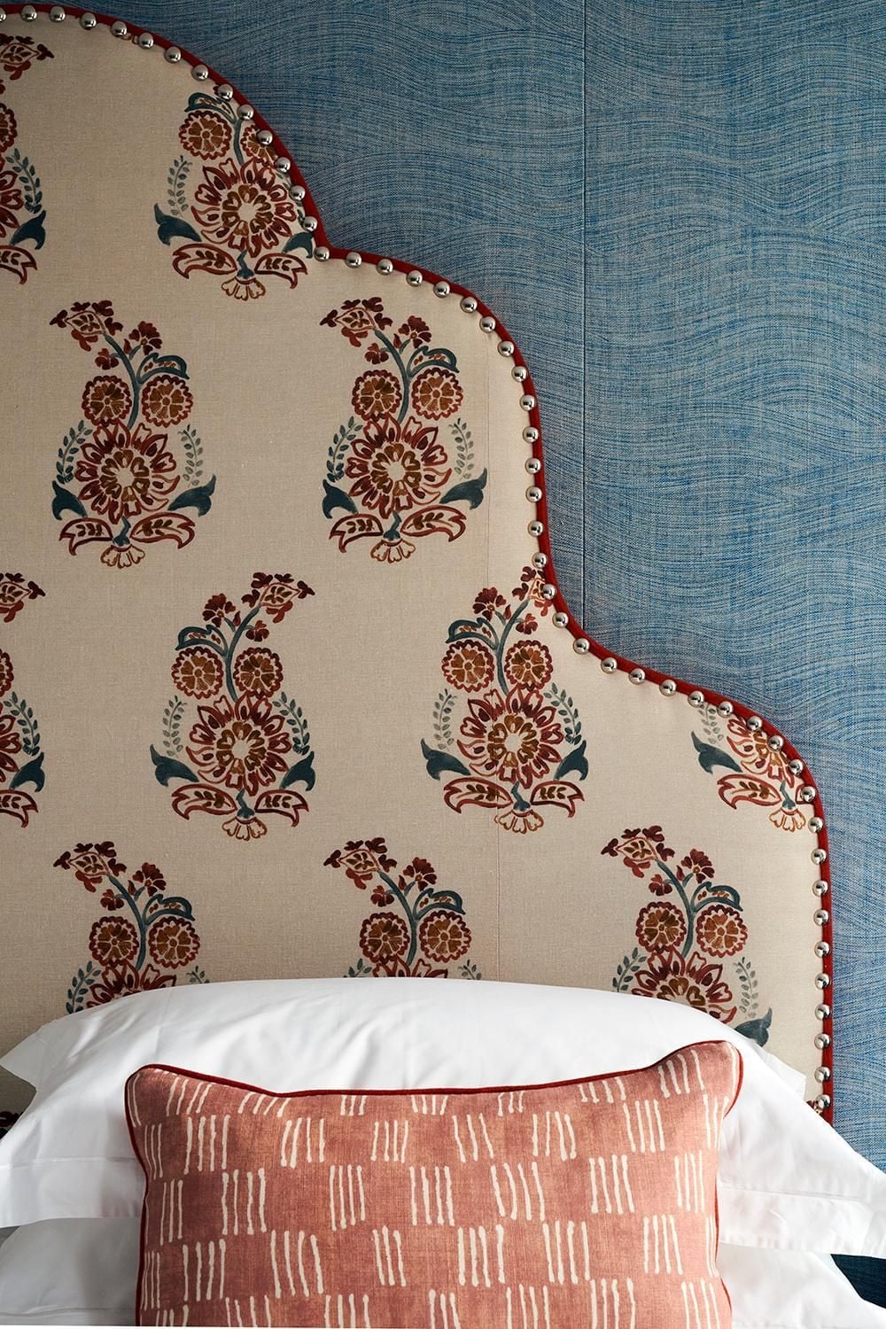 Hand-blocked florals and nailhead trim create an intimate headboard moment, while coral-toned linens add warmth against textured blue wallcovering in this thoughtfully layered guest room.
