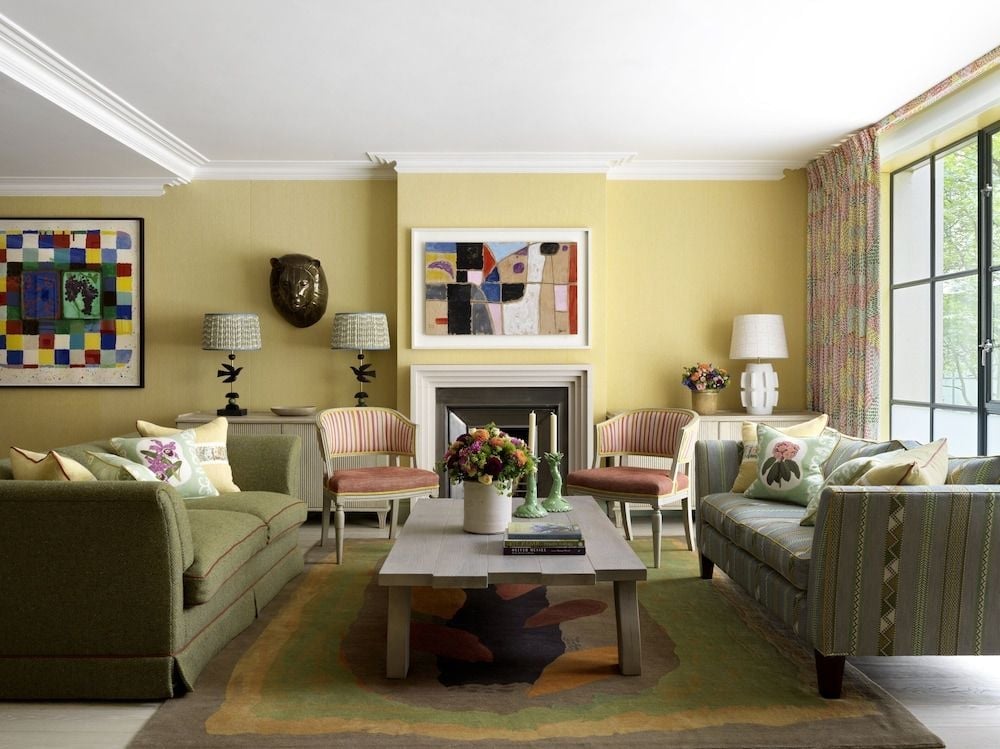 Sage green sofas and cheerful yellow walls create an inviting living space, where contemporary art mingles with striped armchairs beside a crackling fireplace.