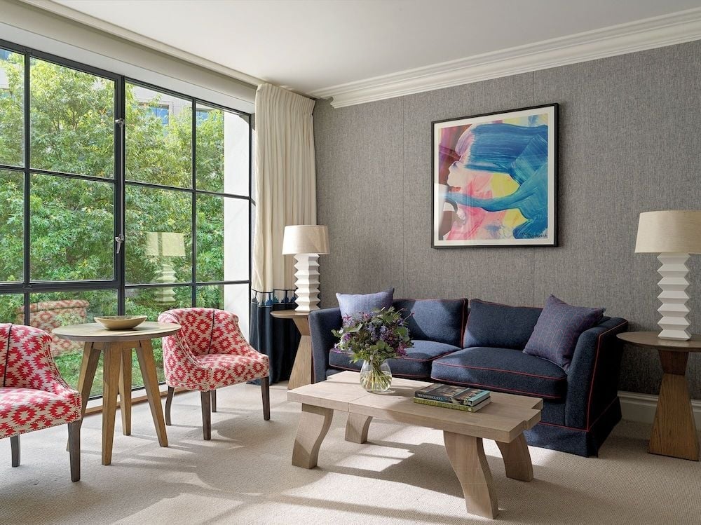 Sunlight streams through industrial-framed windows onto plush navy sofas and crimson geometric chairs, creating an intimate London retreat where contemporary art meets sophisticated comfort.