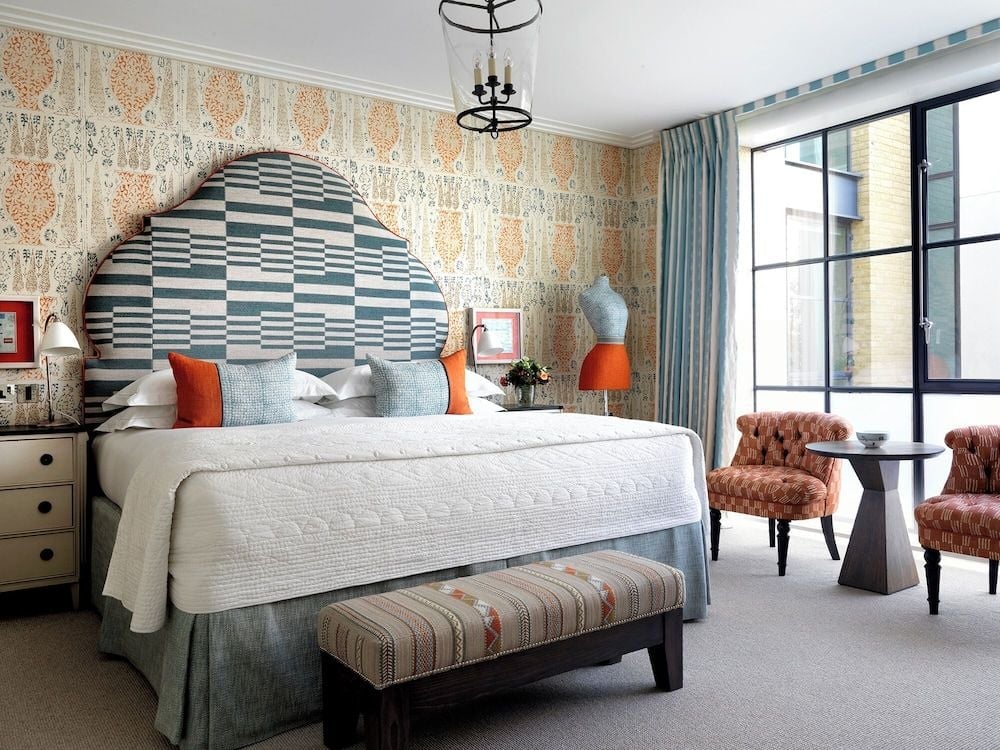 Geometric headboards and tangerine accents create playful sophistication in this Soho sanctuary, where patterned wallpaper meets plush velvet seating beside floor-to-ceiling windows.