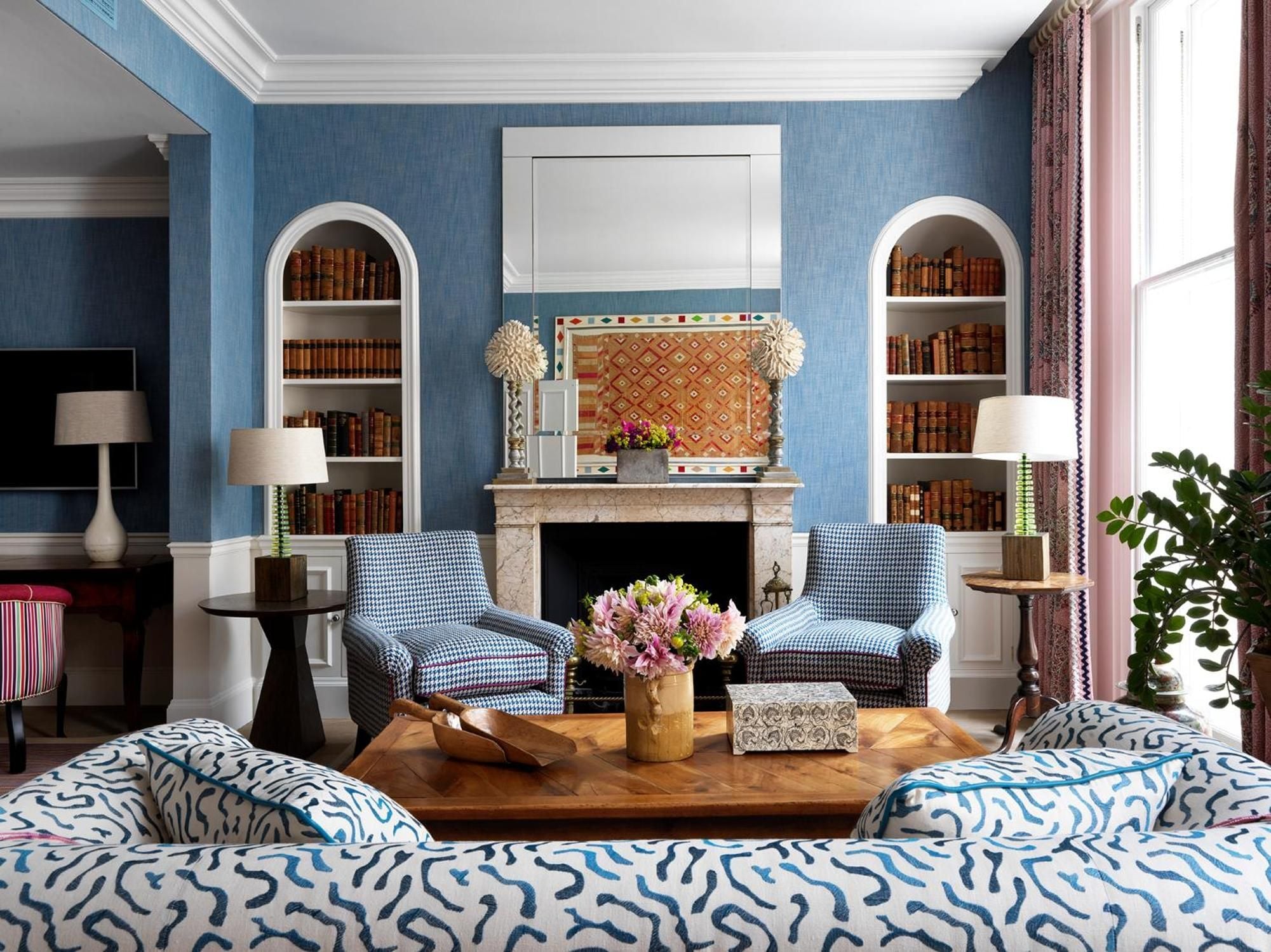 Gingham armchairs and botanical patterns create intimate conversation nooks around the fireplace, where leather-bound volumes line arched alcoves in this sophisticated London retreat.