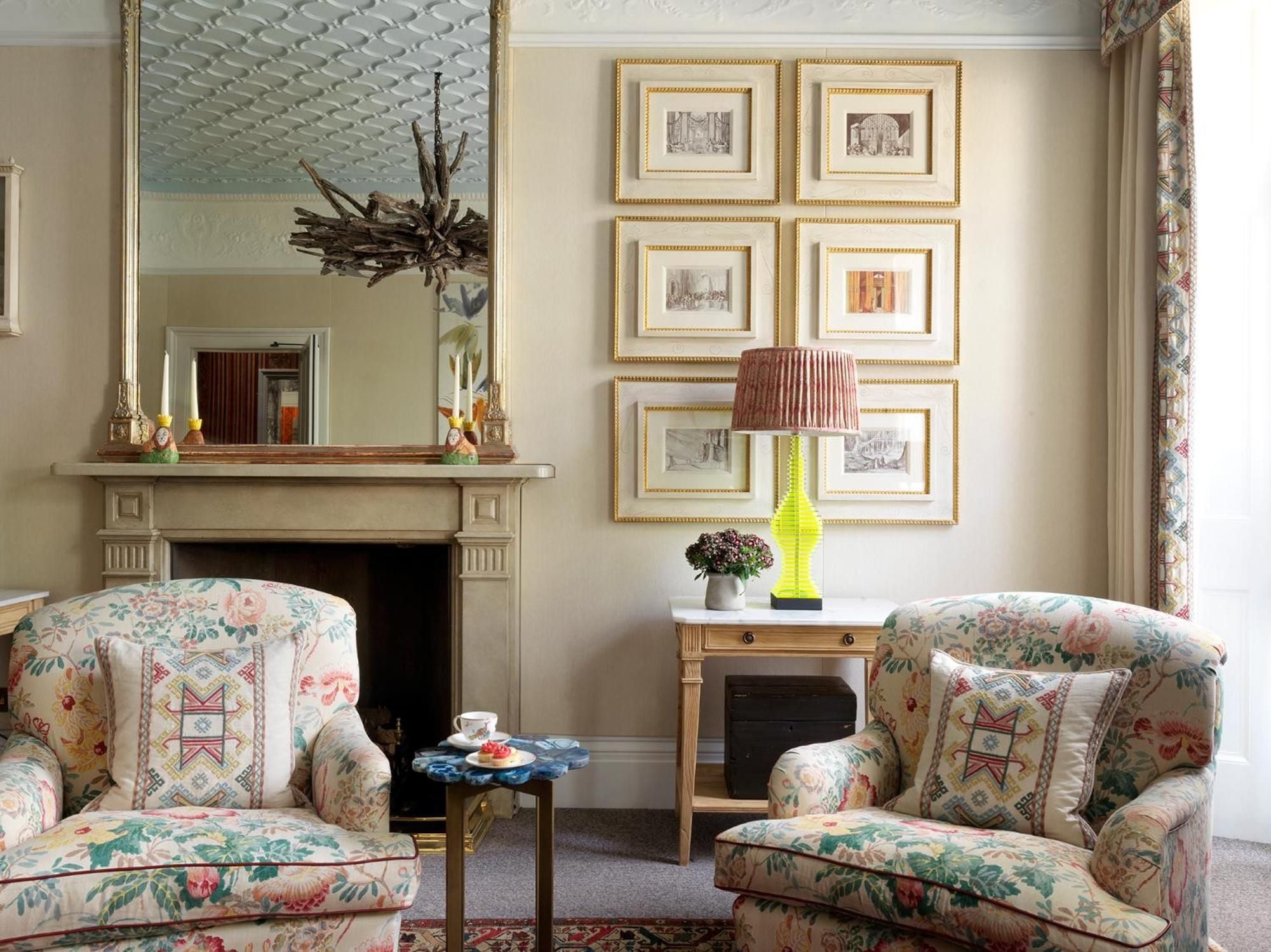 Floral armchairs frame the fireplace while gilt-framed prints create an intimate gallery wall, epitomizing the boutique hotel's refined English country house charm.