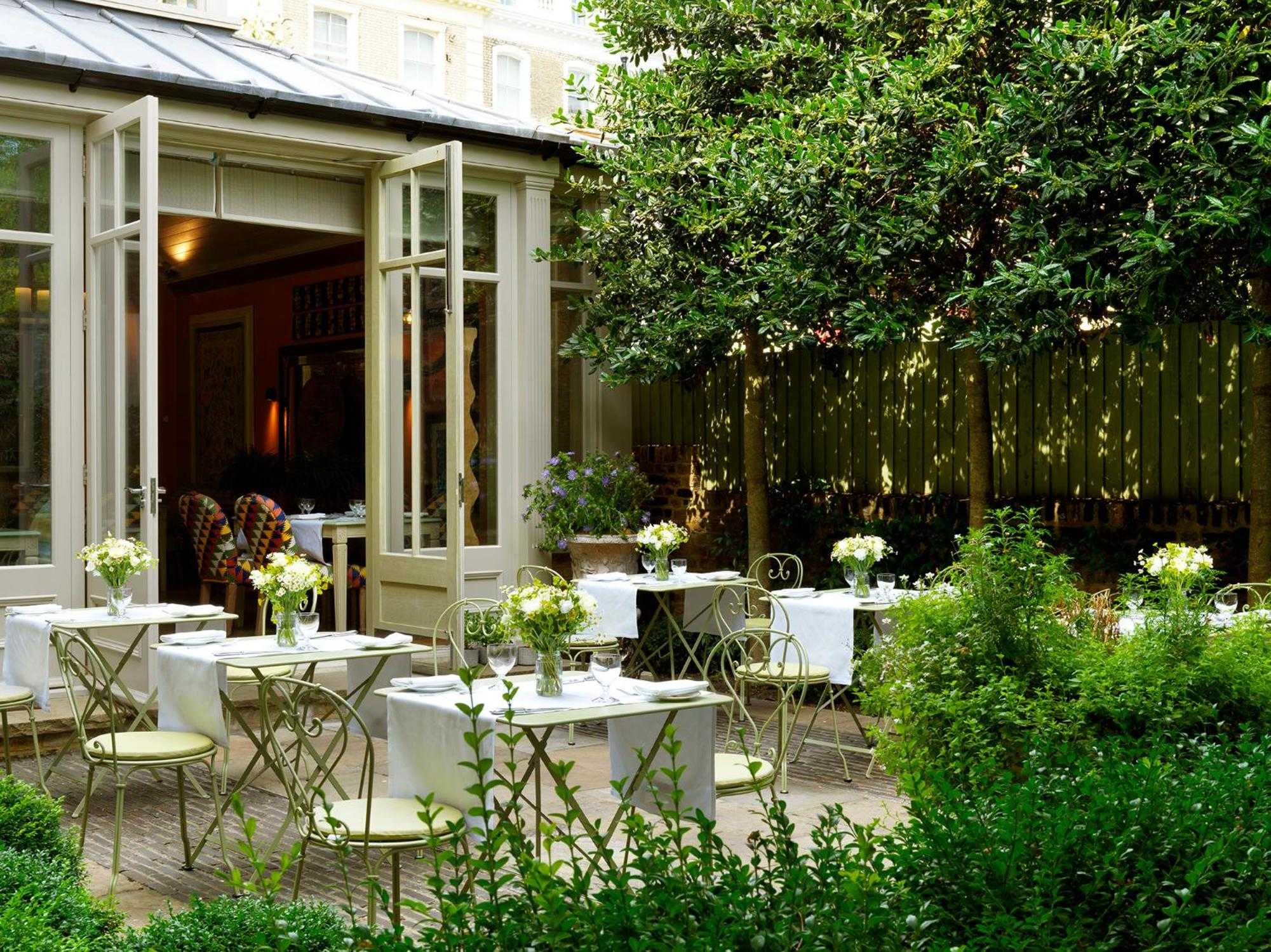 White-linen tables nestled among emerald foliage create an intimate courtyard dining sanctuary, where dappled sunlight filters through towering trees onto weathered stone.