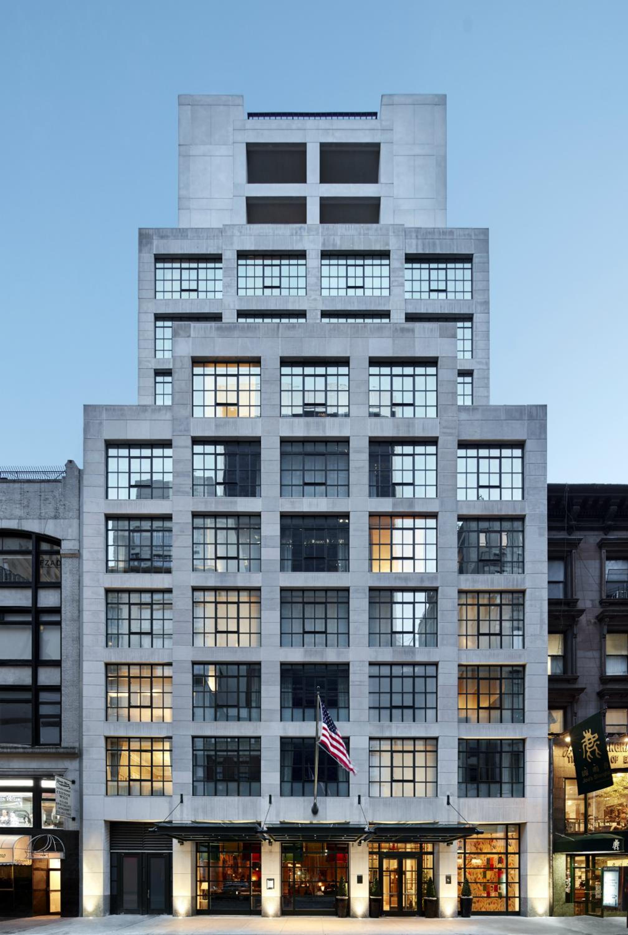 Soaring limestone façade rises like a modern fortress above SoHo's cobblestones, its geometric windows glowing warmly against the evening sky while the American flag marks this boutique sanctuary.