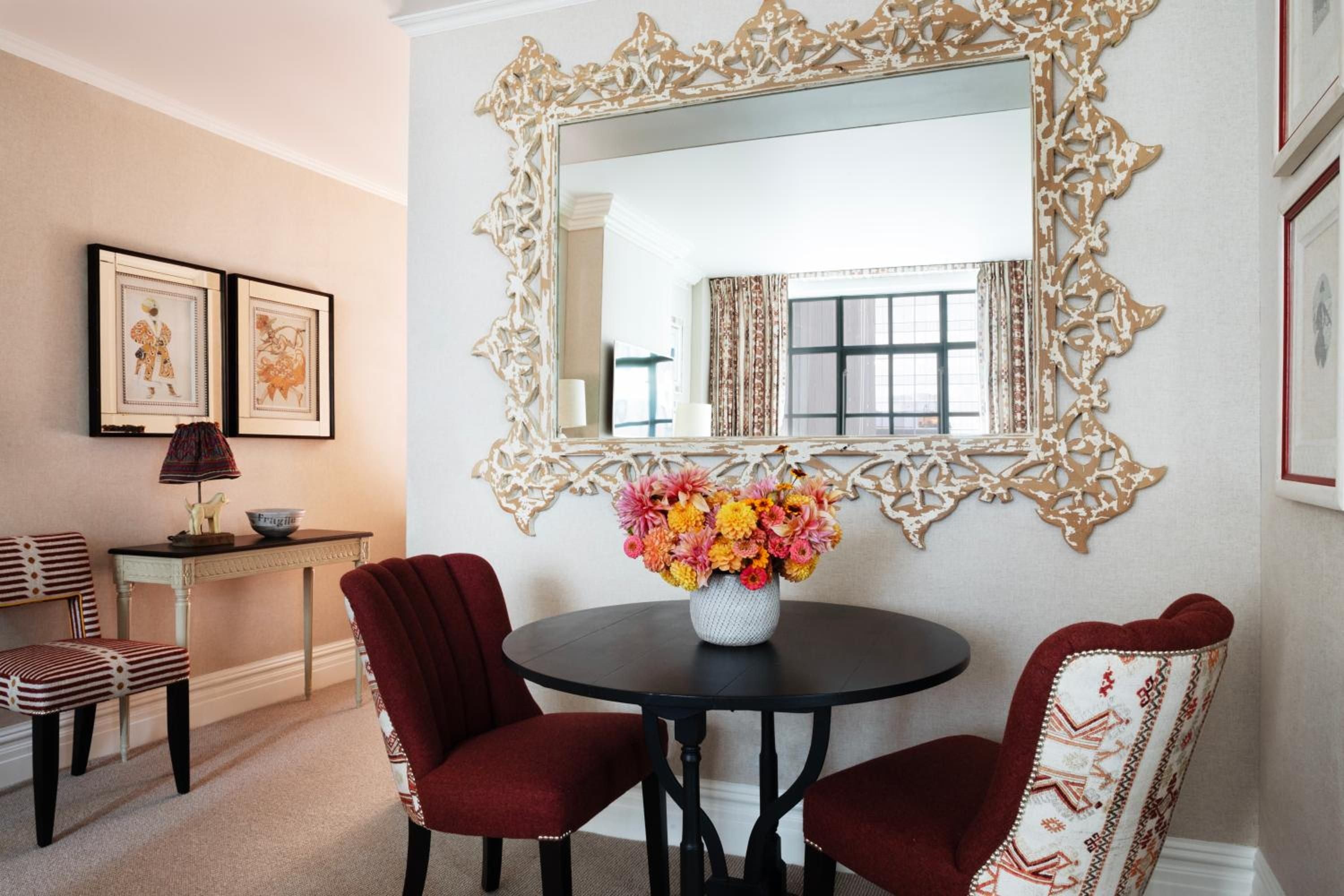 Ornate gold mirrors and jewel-toned velvet chairs create an intimate dining corner where afternoon light filters through patterned curtains, crowned by vibrant dahlias.