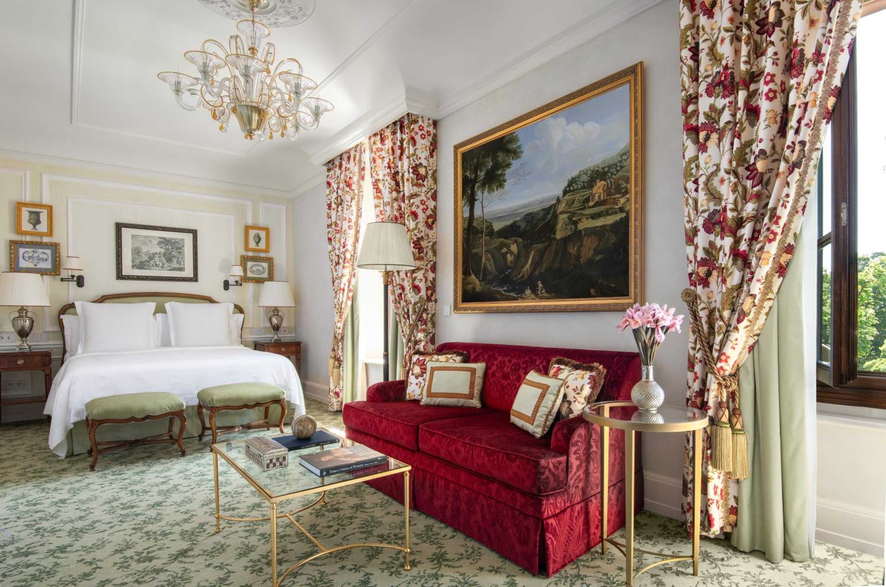 Venetian chandeliers cast gentle light over sumptuous red velvet seating and botanical curtains, while Renaissance landscapes adorn walls in this palatial Florence suite.