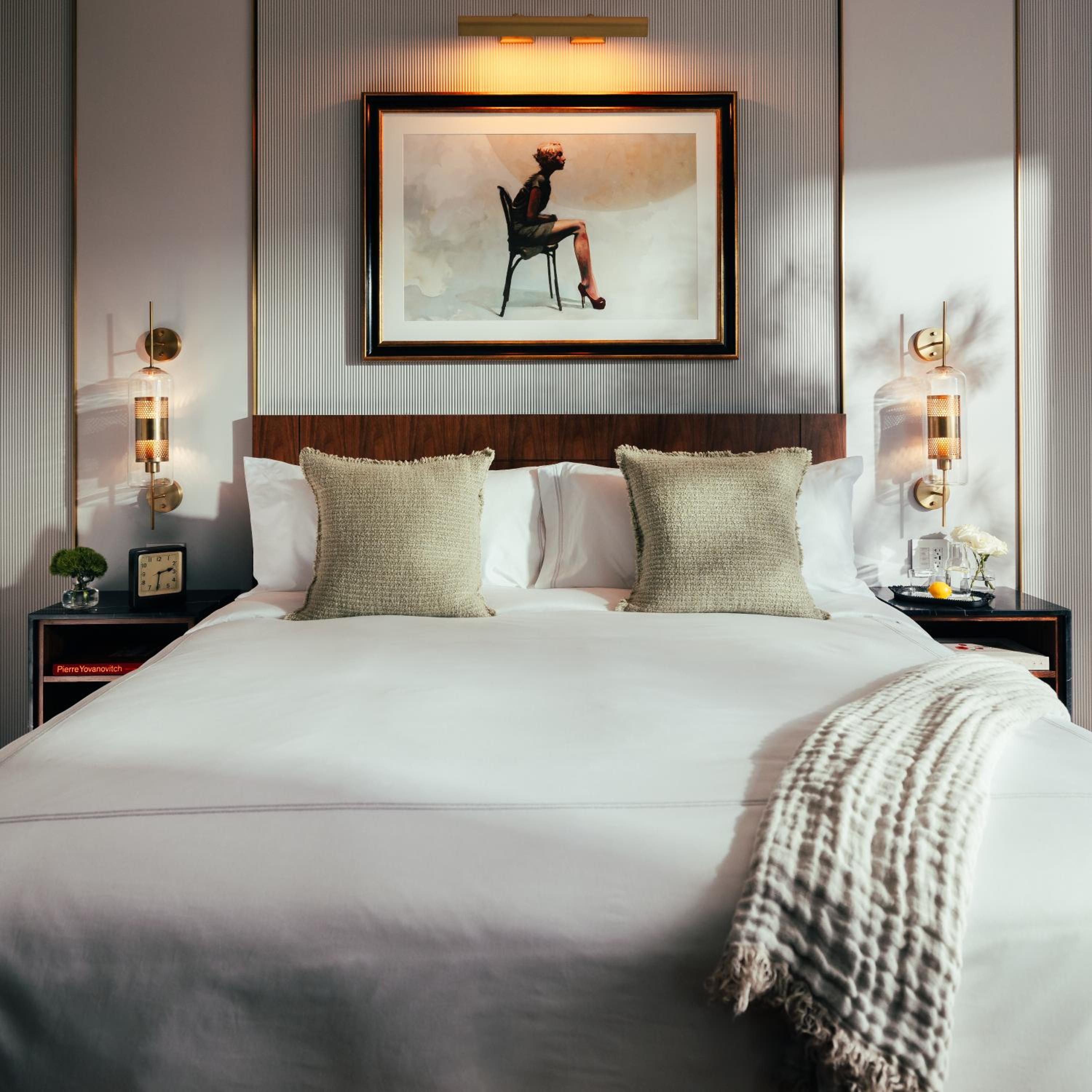 Crisp white linens meet warm walnut headboards beneath carefully curated artwork, while brass sconces cast an intimate glow across textured pillows and ribbed wall panels.