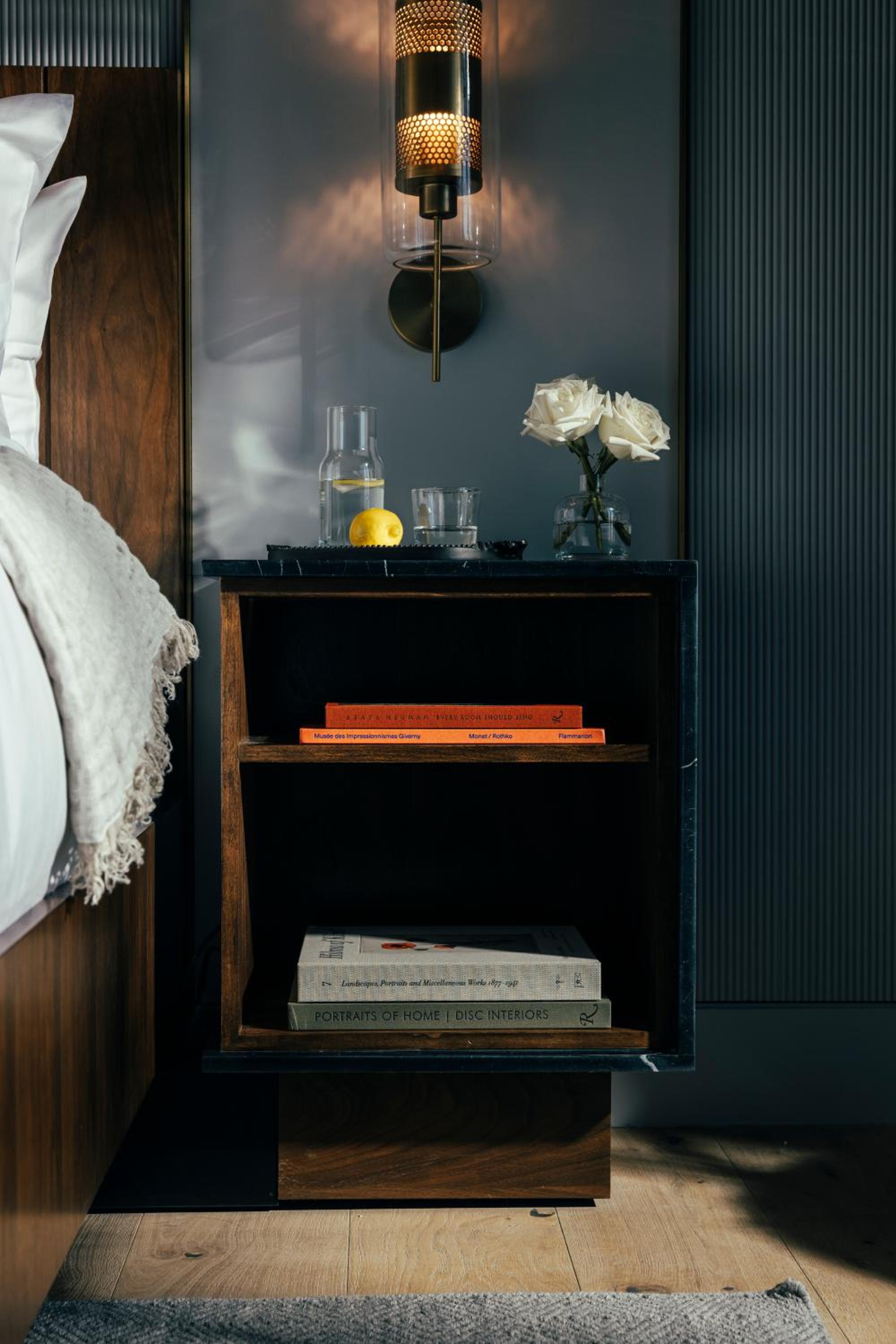 Warm brass sconces cast honeyed light across the thoughtfully curated bedside vignette, where crisp white roses and carefully chosen books create an intimate reading retreat.